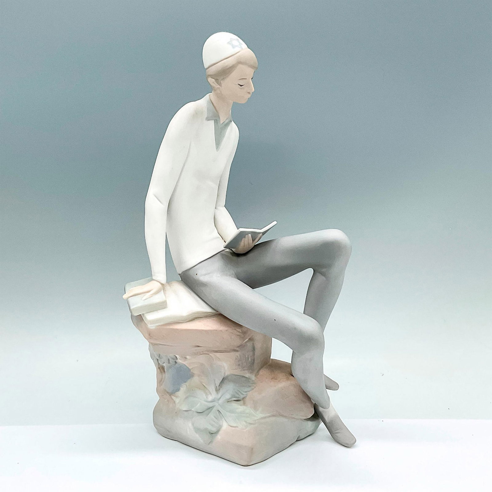 Hebrew Student 1014684 - Lladro Porcelain Figurine: A matte figure of a young man seated on a boulder reading. Lladro backstamp. Artist: Juan Huerta Issued: c. 1970-1985 Dimensions: 6"L x 4.5"W x 11.5"H Manufacturer: Lladro Country of Origin: Spai