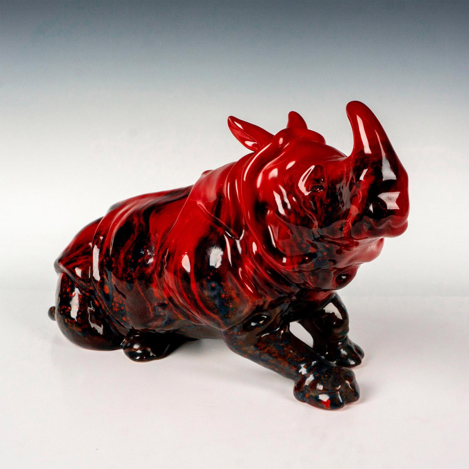 Royal Doulton Flambe Veined Sculpture, Rhino Lying: A red glazed seated rhinoceros with dark blue blemishes. Royal Doulton Flambe Veined backstamp. Artist: Leslie Harradine Issued: c. 1973-1996 Dimensions: 15.5"L x 6.75"W x 9.5"H Manufacturer: Royal Do