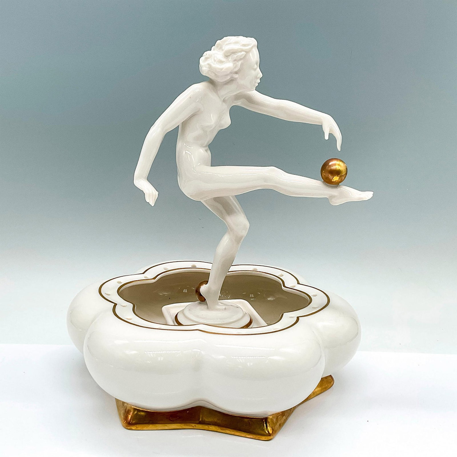 Hutschenreuther Art Deco Figurine with Base, Sun Child: A white nude figure, also known as Sun Worshipper. She is balancing a gold orb on her leg. Incudes a floral shaped white bowl base with gilded accents. Hutschenreuther backstamps. C. Werner marked on 