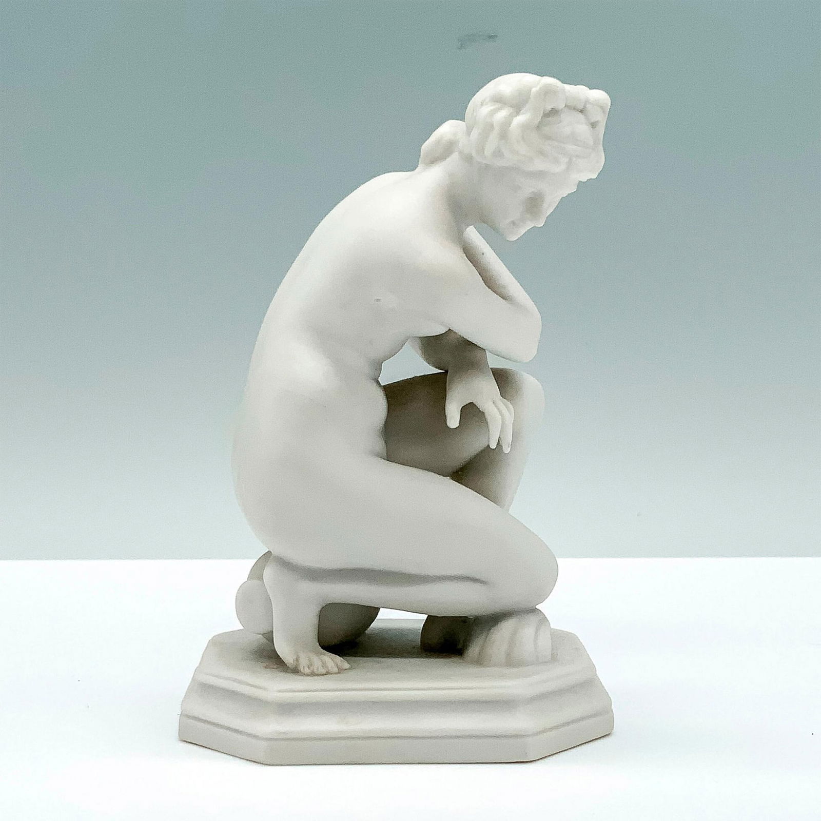 Antique Scheibe-Alsbach Parian Crouching Venus Figurine: A white figurine made as the crouching Venus with one knee on the ground and one arm covering the subject's breasts while she sits on a large vase. Marked on the bottom. Dimensions: 3"L x 2.25"W x 4.2