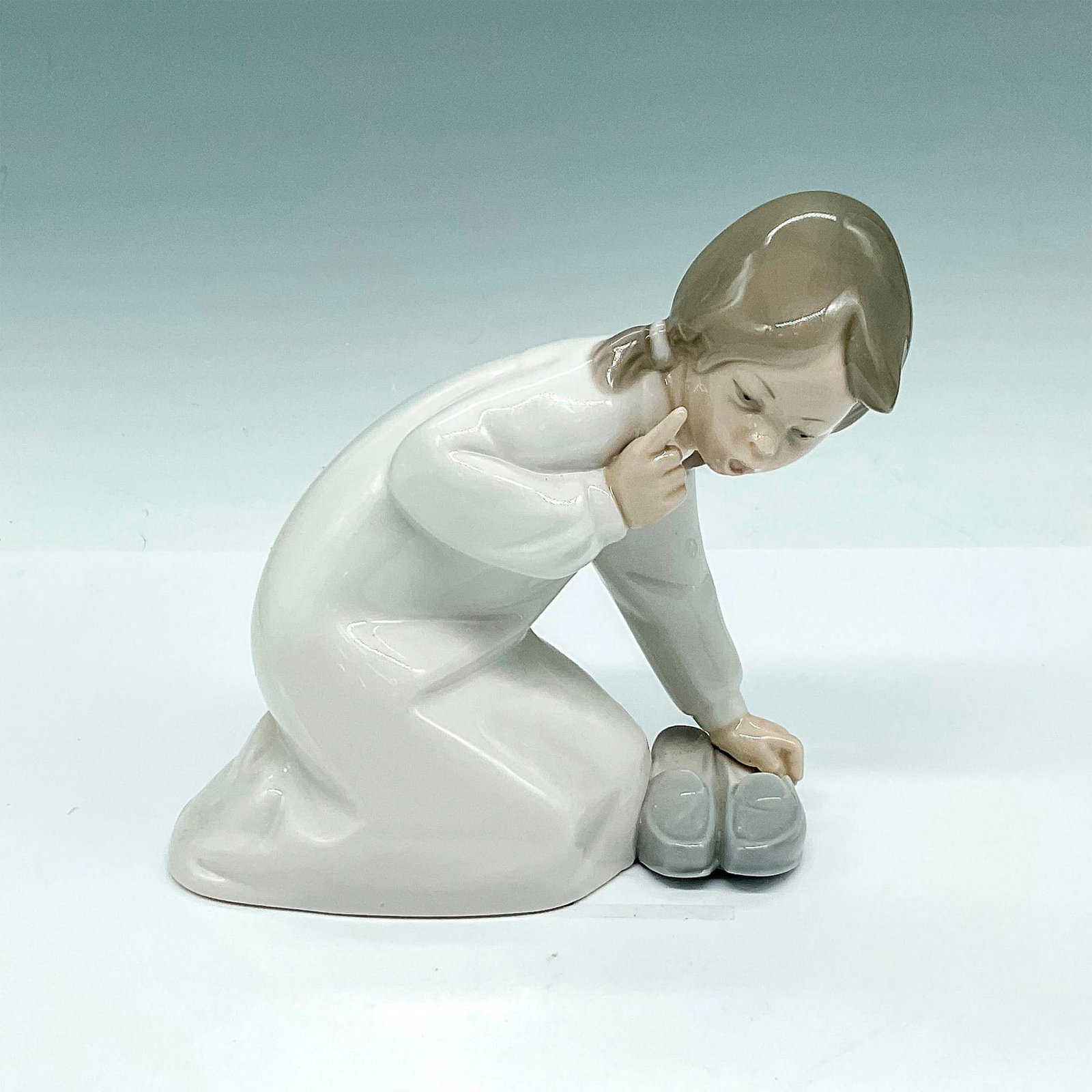 Little Girl with Slippers 1004523 - Lladro Porcelain Figurine: Glossy finish figurine of little girl crouching down to pick up slippers. Lladro backstamp. Artist: Vincente Martinez Issued: 1969-1993 Dimensions: 5.75"L x 3"W x 5.5"H Manufacturer: Lladro Country o
