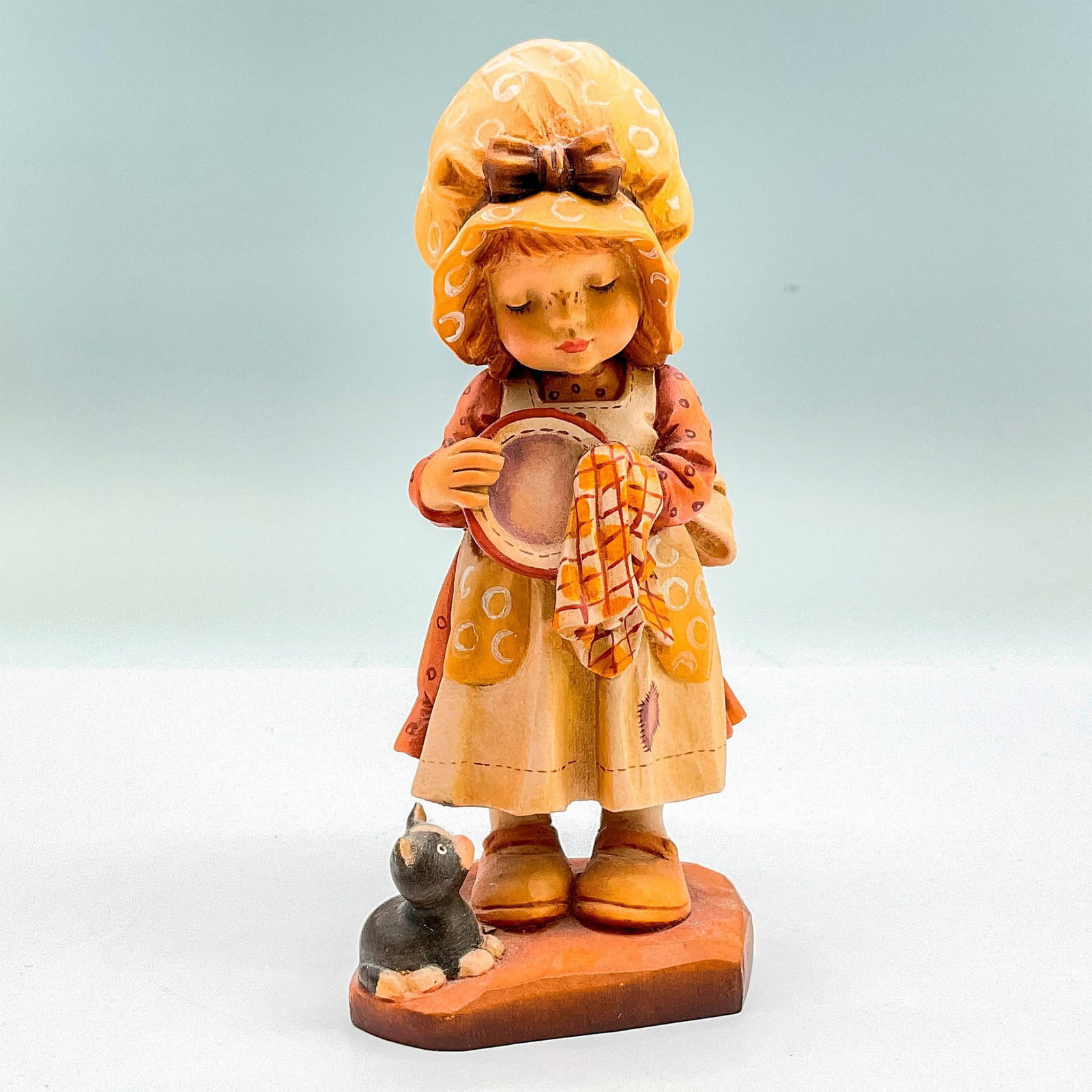 Anri Italy Wood Carved Figurine, Helping Mother: Cheerful painted figure of a young girl drying a plate while her little cat looks up at her. In hues of earthy orange, yellow, tan and brown. Anri metal logo emblem affixed to base. Design Valentine b