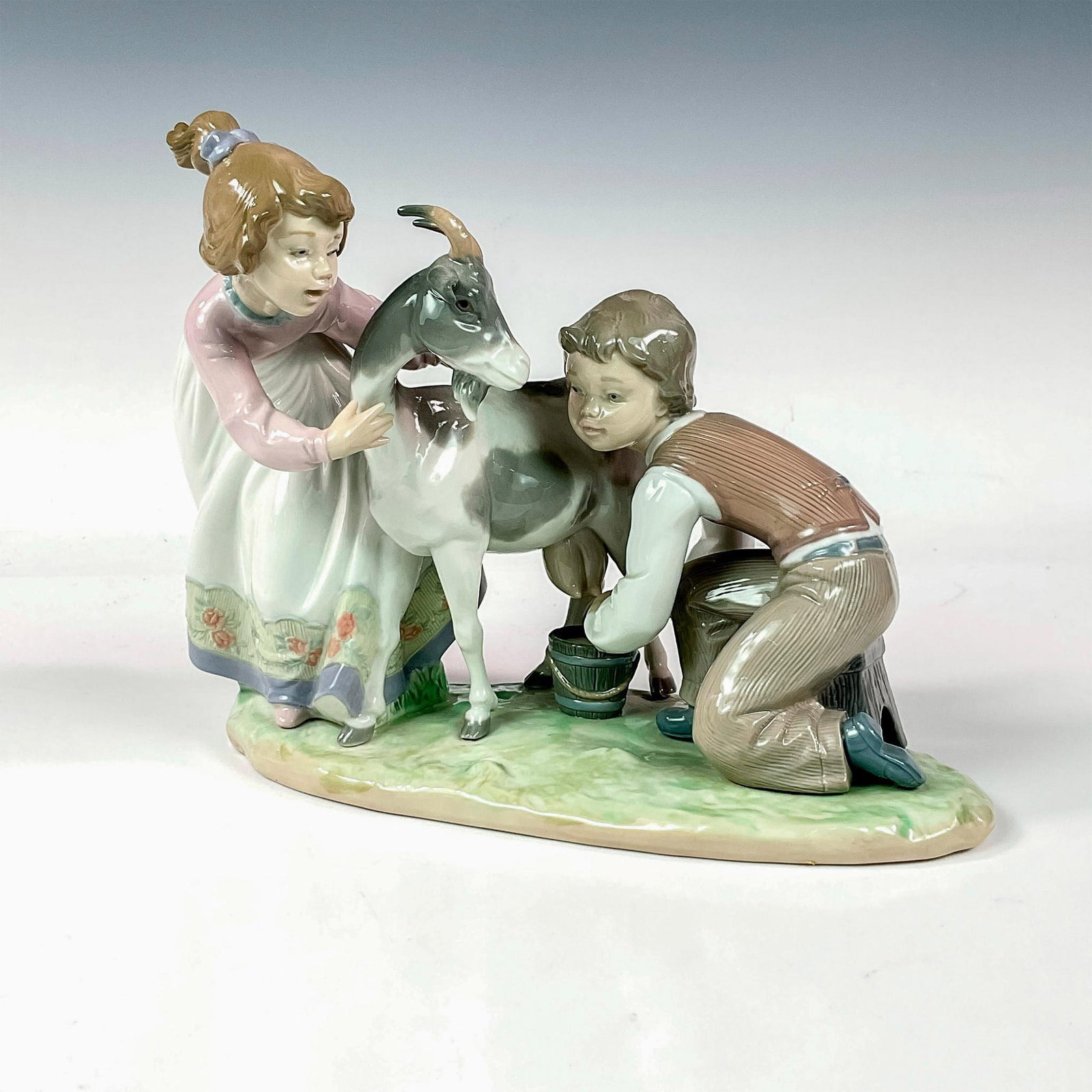 Hold Her Still 1005753 - Lladro Porcelain Figurine: Glossy finish, modeled as a girl and boy milking a goat. Lladro backstamp. Artist: Regino Torrijos Issued: 1991-1993 Dimensions: 10.25"L x 4.25"W x 8"H Manufacturer: Lladro Country of Origin: Spa