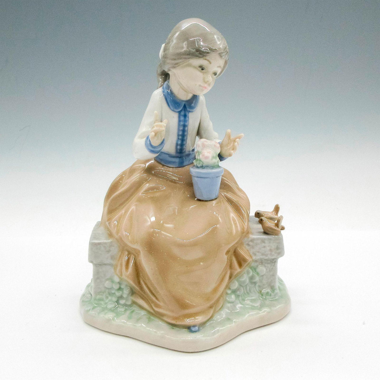 Nao by Lladro Porcelain Figurine, Sitting on Garden Bench: Glossy figural with birds and flowers. Tan and blue coloration. Nao by Lladro backstamp. This item has its original box: 9"L x 7.5"W x 6.5"H. Issued: 20th century Dimensions: 5"L x 3.5"W x 7"H Manufac