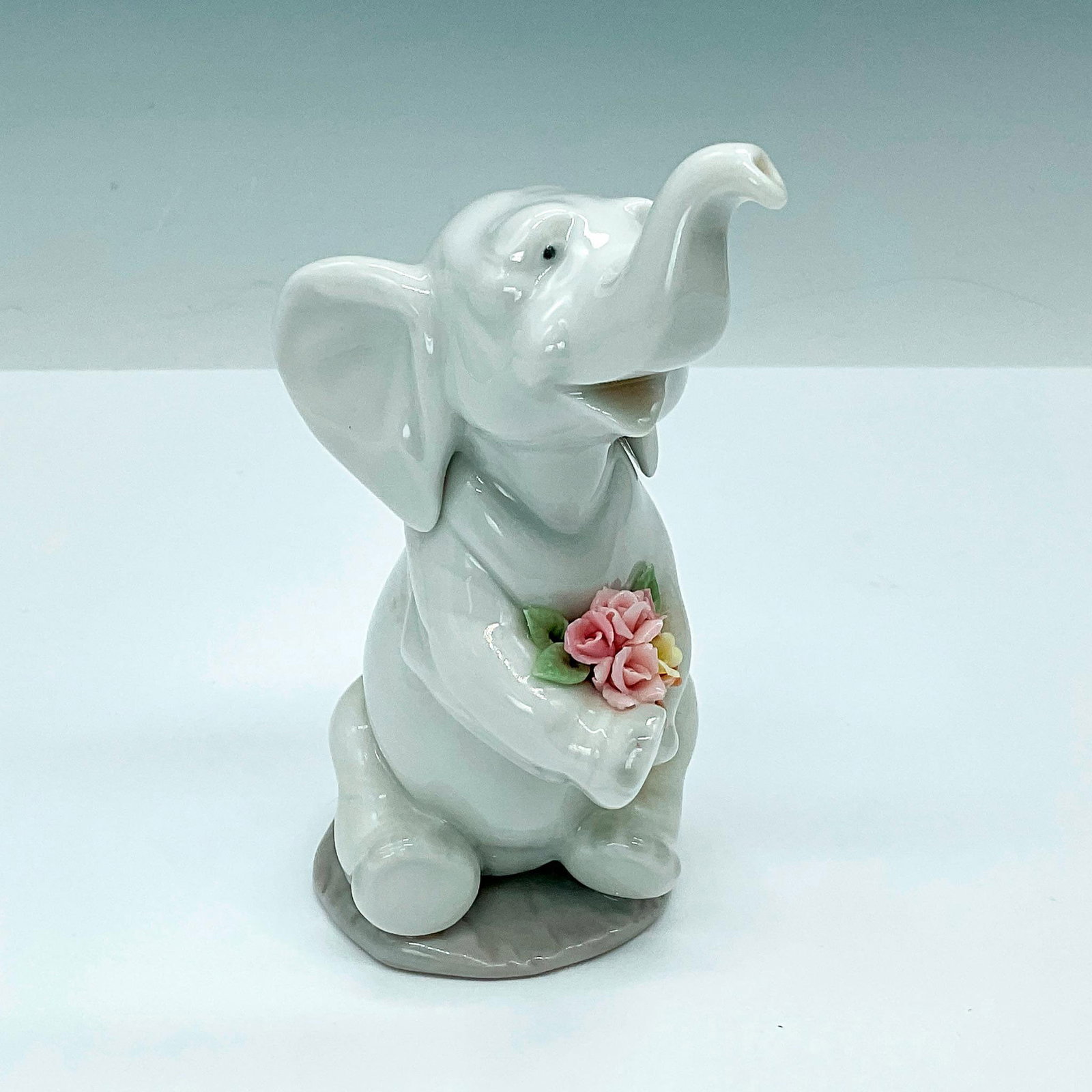 Lucky in Love 1006462 - Lladro Porcelain Figurine: A cute figurine featuring a white elephant sitting and delicately holding a beautiful bouquet of pink flowers. Lladro backstamp. Issued: 1997-2004 Dimensions: 3.5"L x 2.25"W x 5"H Manufacturer: Lladro