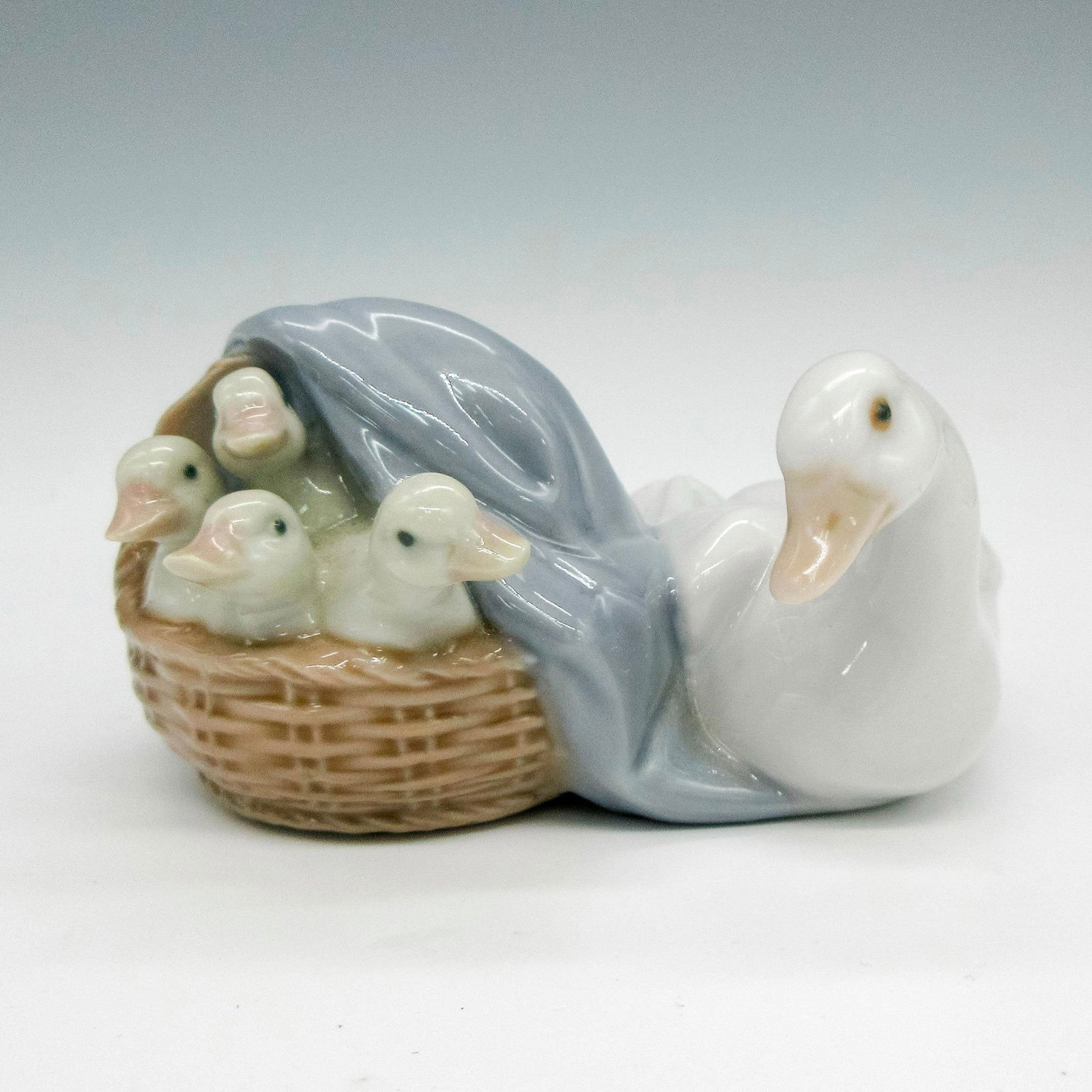 Ducks 1004895 - Lladro Porcelain Figurine: Glazed figural of Momma duck with baby ducks in basket with blue blanket. Lladro backstamp. Issued: c. 1974 Dimensions: 3.75"L x 3.75"W x 2.5"H Manufacturer: Lladro Country of Origin: Spain