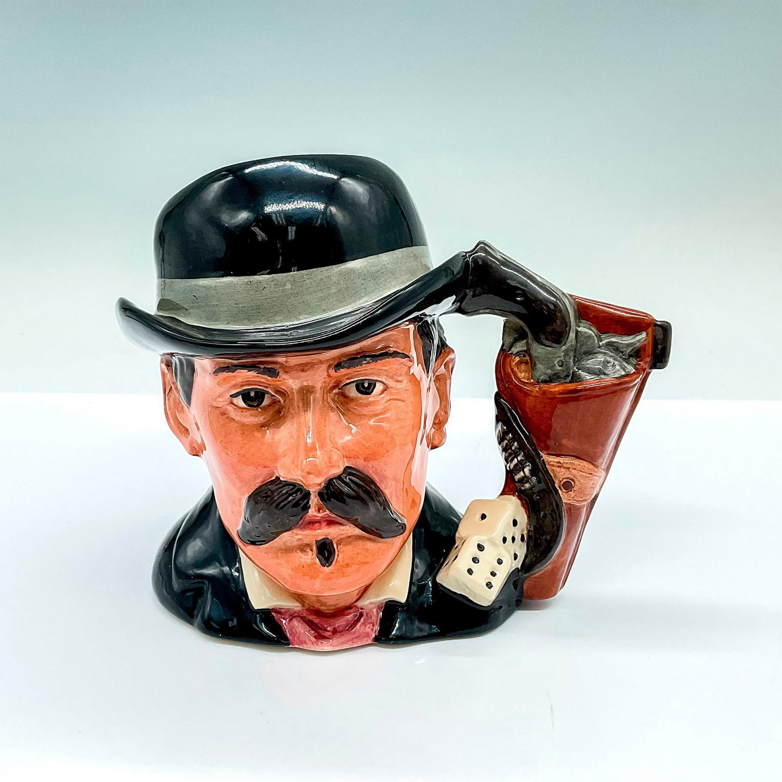Doc Holiday D6731 - Royal Doulton Odd Character Jug: Part of the Wild West Collection; black and gray hat, and black coat; pistol in holster and dice handle. Royal Doulton backstamp. Artist: Stanley James Taylor Issued: c. 1985-1989 Dimensions: 6.5"L x 
