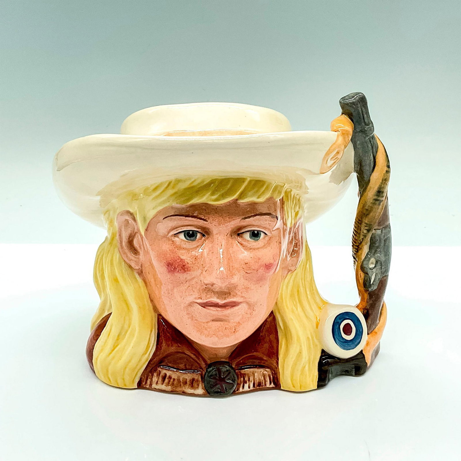 Annie Oakley D6732 - Royal Doulton Odd Character Jug: Part of the Wild West Collection; yellow hair, cream hat, and brown shirt; rifle and belt handle. Royal Doulton backstamp. Artist: Stanley James Taylor Issued: c. 1985-1989 Dimensions: 6.5"L x 5.25"W 