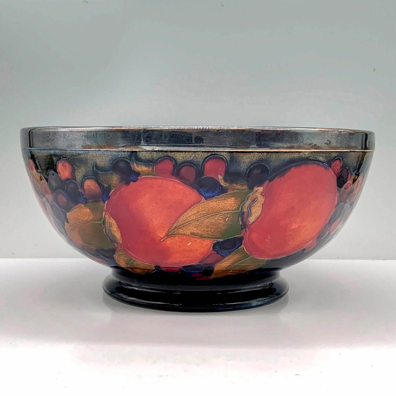 Moorcroft Pomegranate Bowl with Sterling Silver Rim: A blue painted bowl decorated with pomegranates and berries painted throughout with a hallmarked sterling silver rim. Moorcroft backstamp. Artist signed. Artist: William Moorcroft Dimensions: 8"dia x 