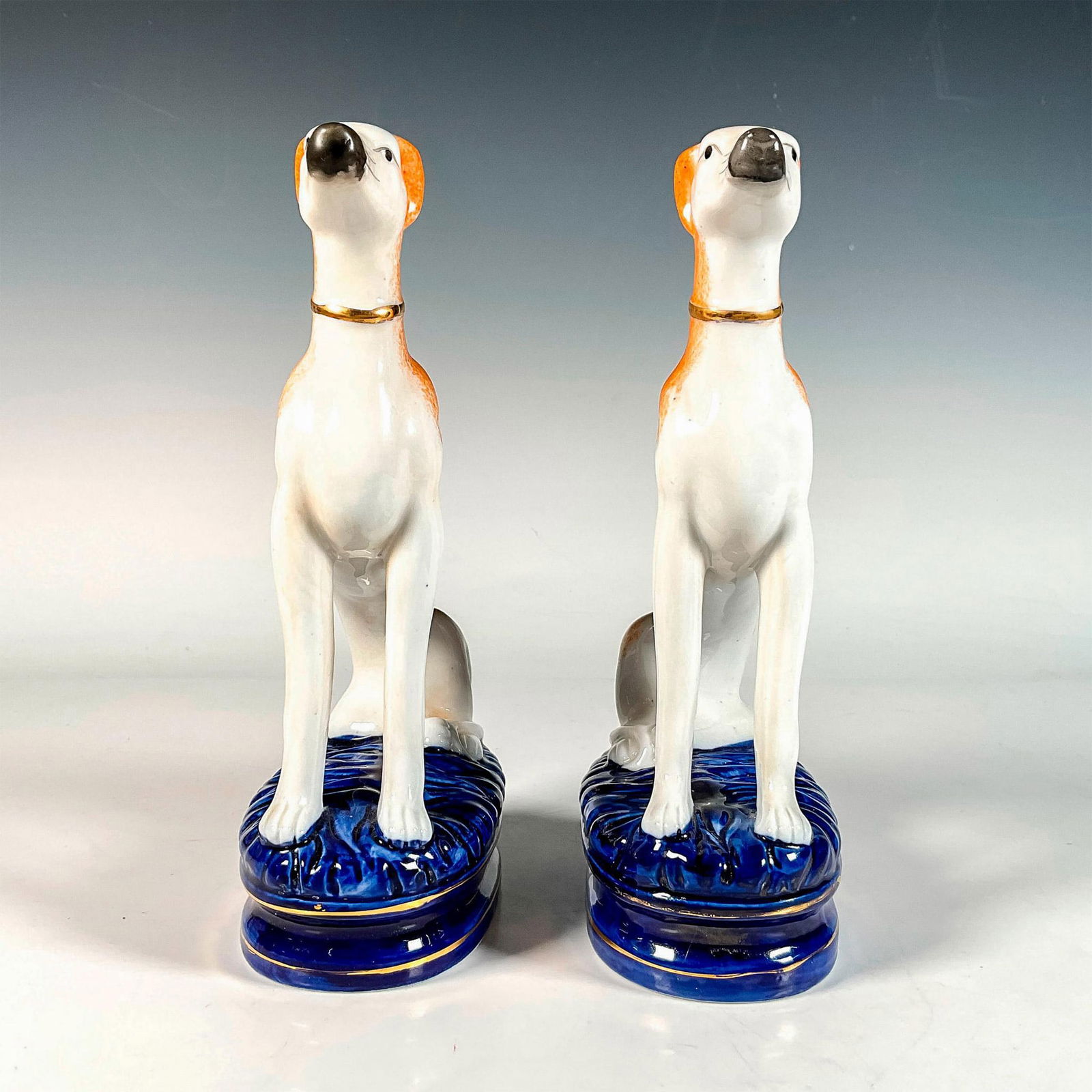 Pair of Staffordshire Pottery Greyhound Bookends: Exquisite figurines depicting sitting greyhounds, painted with an orange coat and gilt collar. They both sit on a blue glaze base. Issued: Late 20th c. Dimensions: 2.5"L x 4.75"W x 9.5"H Country of Or