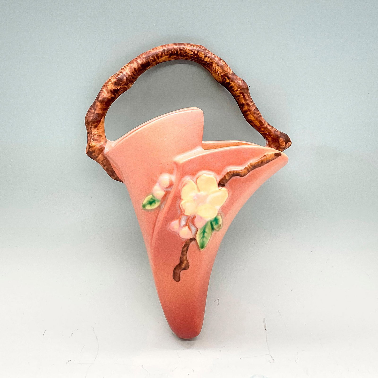 Roseville Pottery Wall Pocket, Apple Blossom: Earthenware wall vase in a dusty pink with a yellow flower and brown top handle. Roseville USA backstamp. Issued: circa 1948 Dimensions: 8.25"H x 6"W x 2.5"D Manufacturer: Roseville Country of Origin: