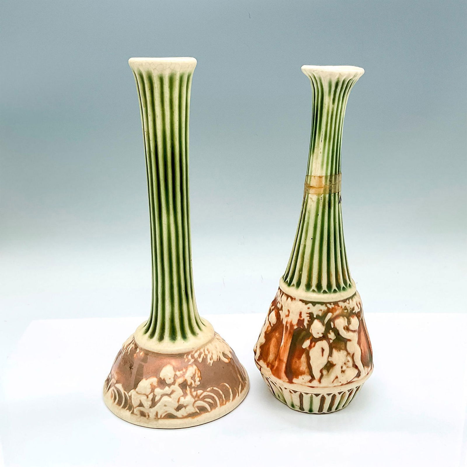 2pc Roseville Pottery Vases, Donatello: 2 slender neck vessels with green and creamed colored Corinthians and cherubs set against brown ground. Vase 1: 4.75"W x 10"H. Vase 2: 3.75"W x 10"H. Artist: Frederick Rhead Issued: c. 1916 Manufactur