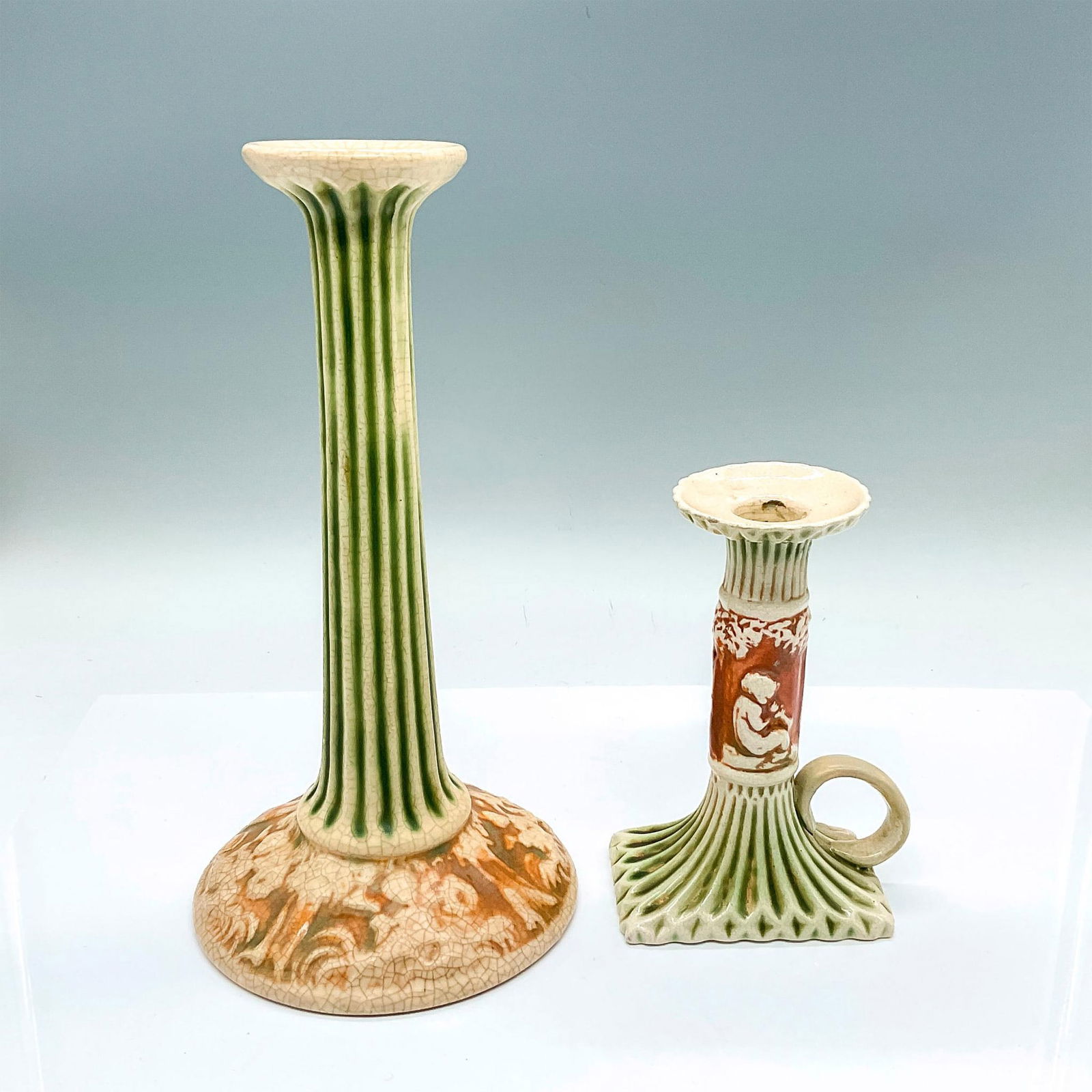 2pc Roseville Pottery Candlestick Holders, Donatello: 2 slender neck candle holders with green and creamed colored Corinthians and cherubs set against brown ground. Candle holder 1: 5.25"W x 10"H. Candle holder 2: 4"W x 6"H. Artist: Frederick Rhead Issue