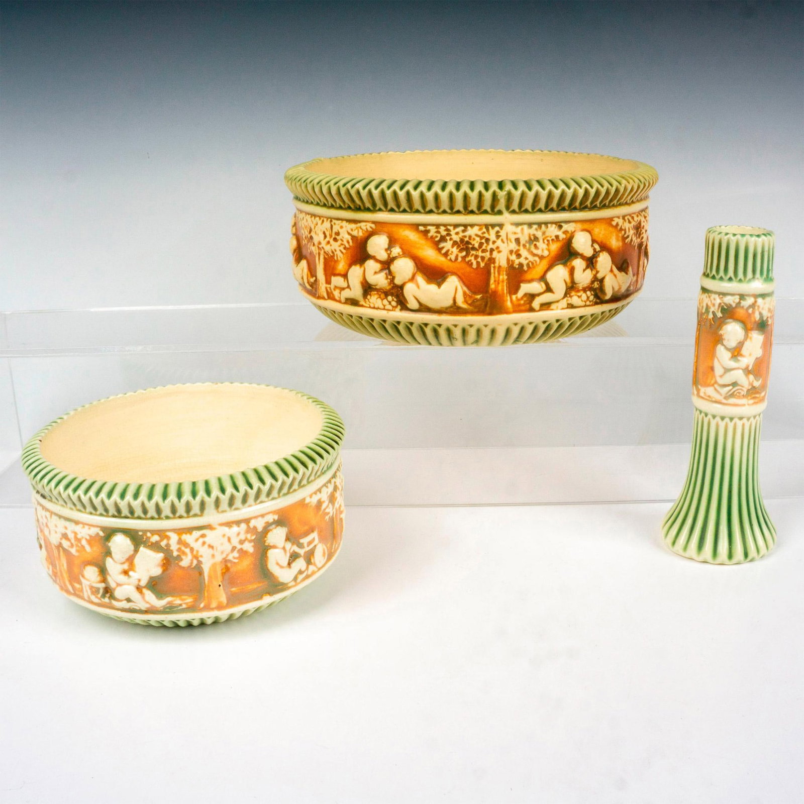 3pc Roseville Pottery Donatello Bowls & Candle Holder: Three pieces with green and brown ground coloration and cream interiors. They are all embossed with cherub scenes. The candle holder measures 2.5"dia. x 6.75"H. The larger bowl measures 8.75"dia. x 3.