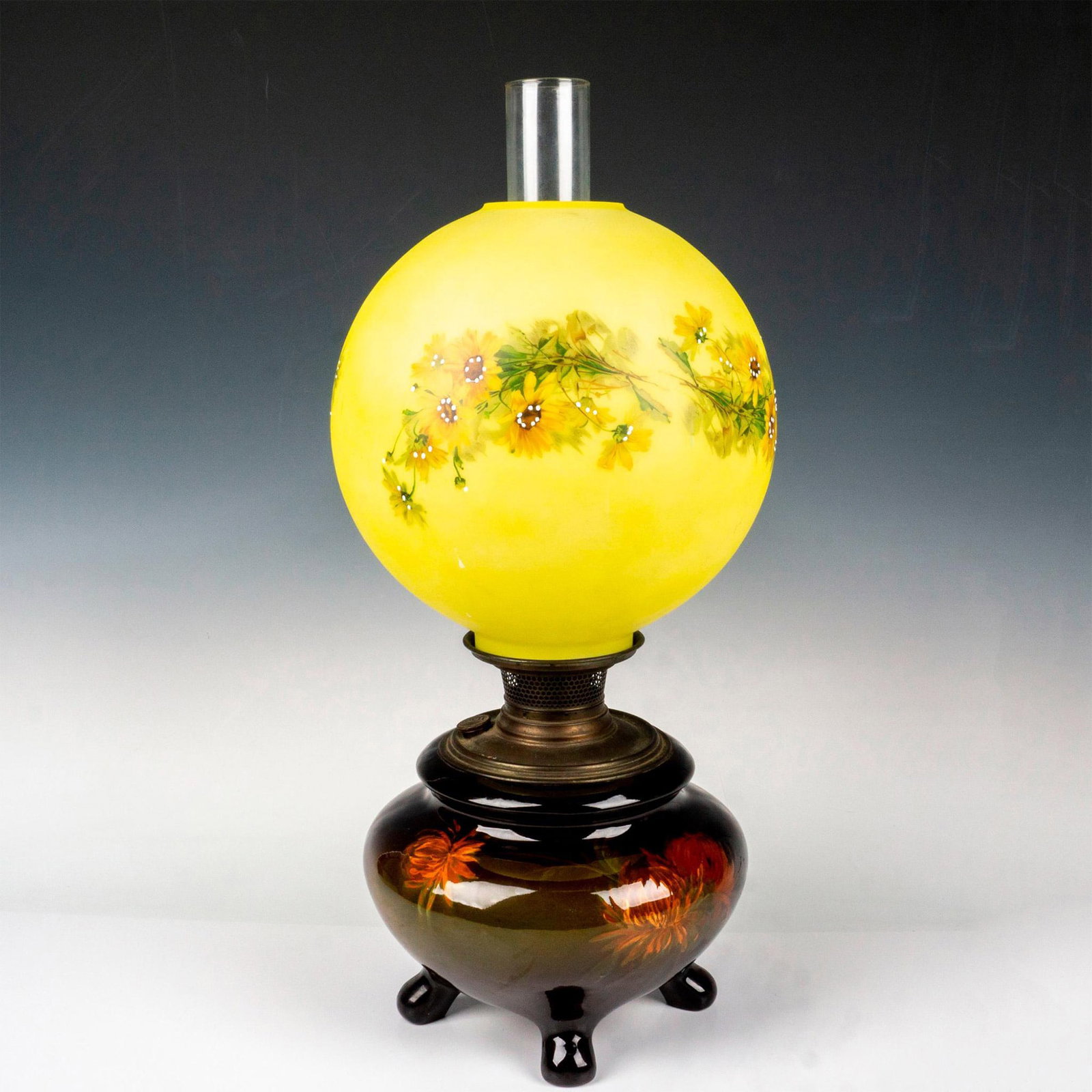 Weller Louwelsa Art Pottery Oil Lamp: Lovely glazed finish featuring a chrysanthemum flower pattern set on a brown-green ground. It has a spectacular form, on four feet with metal hardware. Accompanied by a yellow hurricane glass shade wh