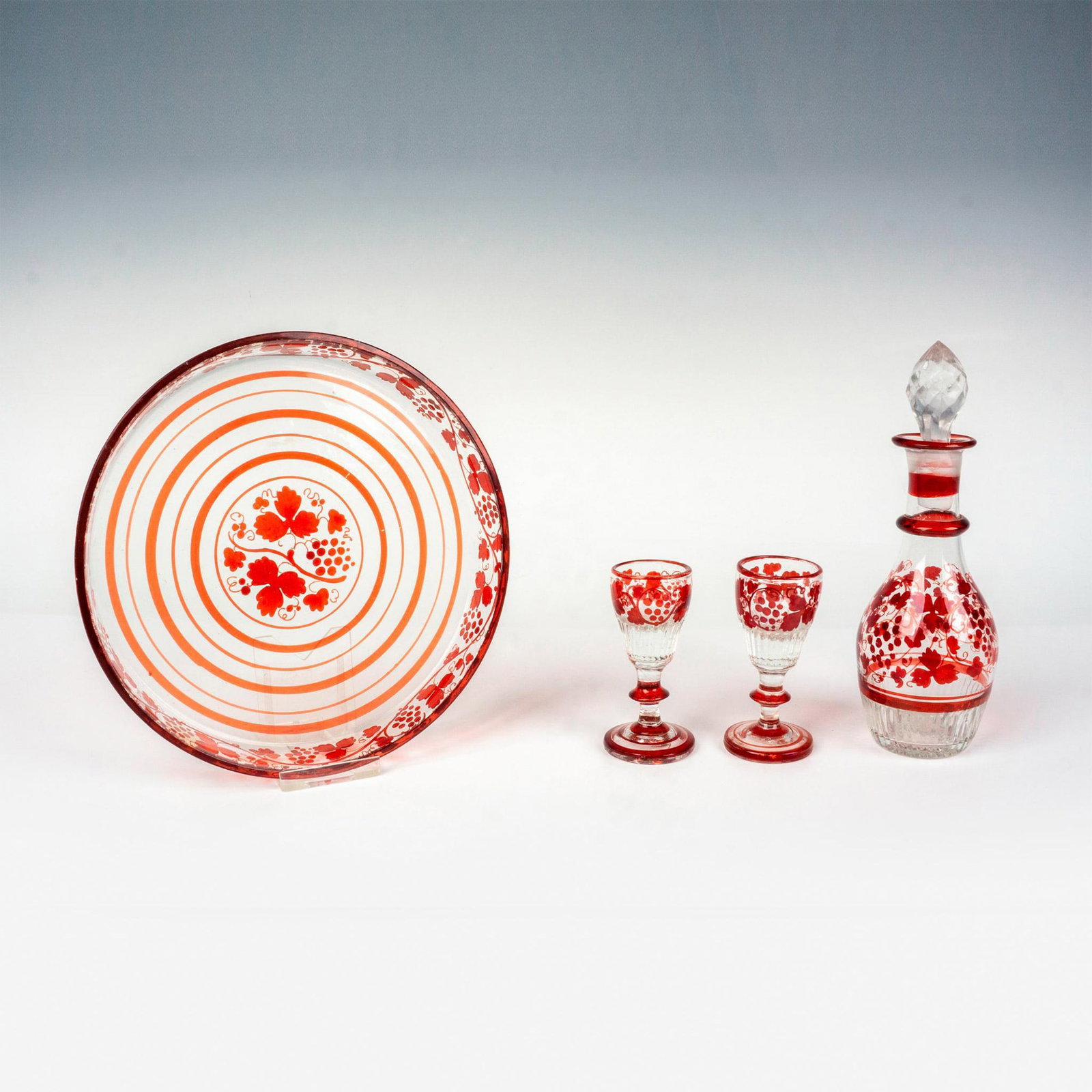 4pc Bohemian Glassware Decorative Set: Ruby red to clear set that includes 2 cordial glasses, 1 decanter with stopper, and 1 tray. This nice set is decorated with grapes and vines. Glasses measure: 1.5"W x 3"H. Decanter measures: 2"W x 6.5