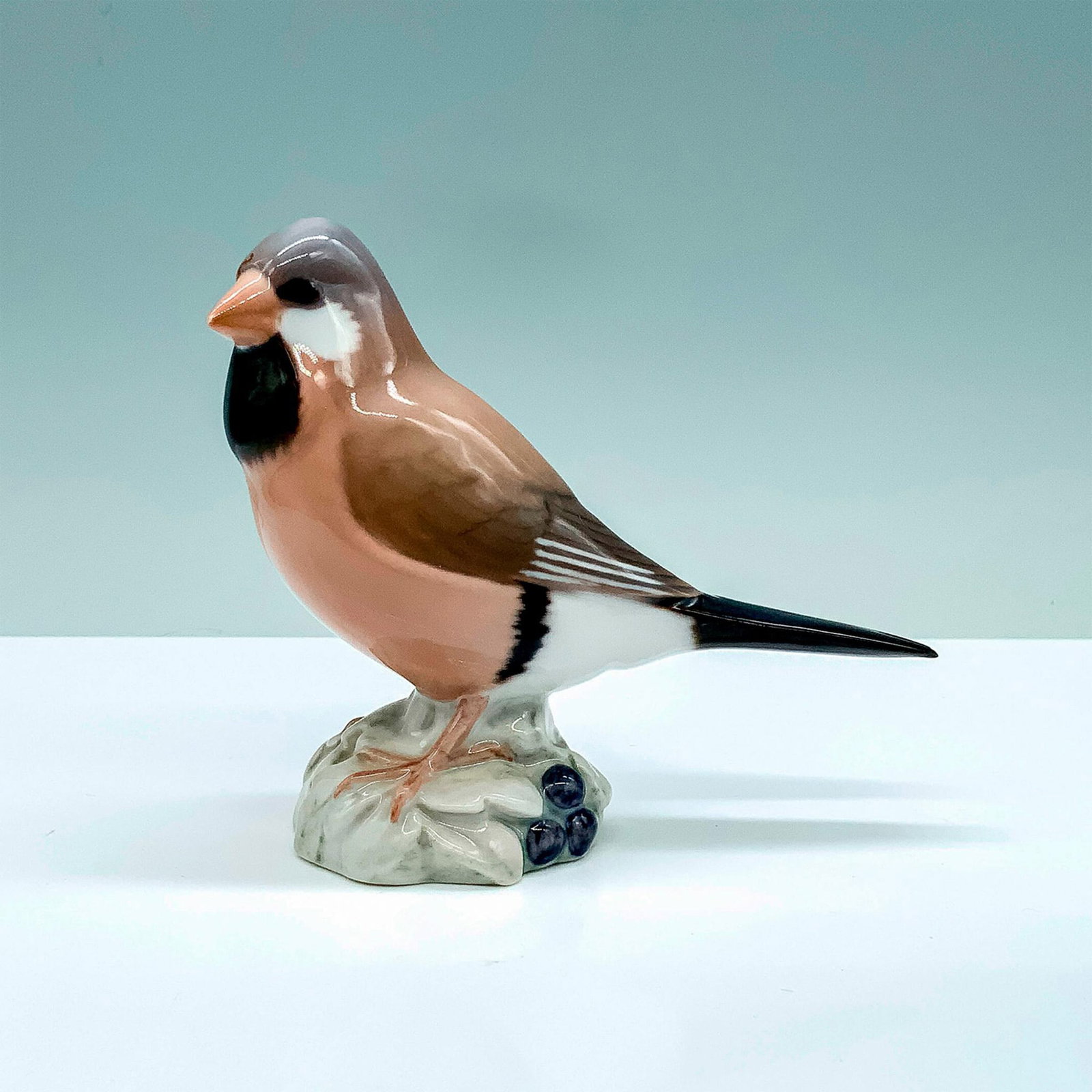 Bing & Grondahl Porcelain Finch Figurine: A figure that depicts a small bird that has a pink, brown, and gray coloration standing atop of a snowy blueberry bush. B and G backstamp. Dimensions: 5"L x 2"W x 4.25"H Manufacturer: B and G Country 