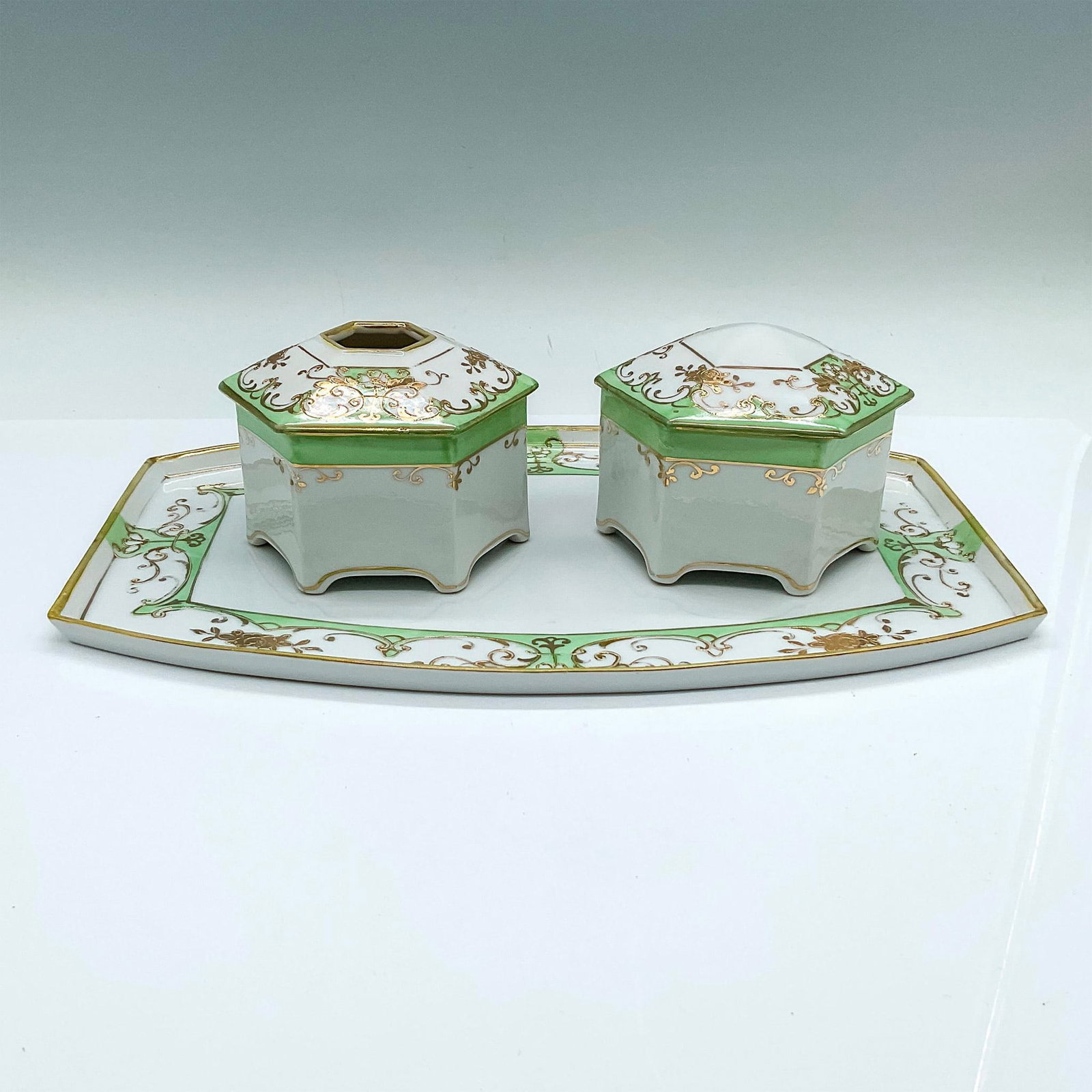 3pc Nippon Porcelain Floral Gold Leaf Vanity Set: A beautiful white set adorned with a light green border and gilt flowers. Includes a tray, hair receiver, and a covered powder box. Nippon backstamps. Tray: 11"L x 7.5"W x 0.5"H. Hair receiver: 4"L x 
