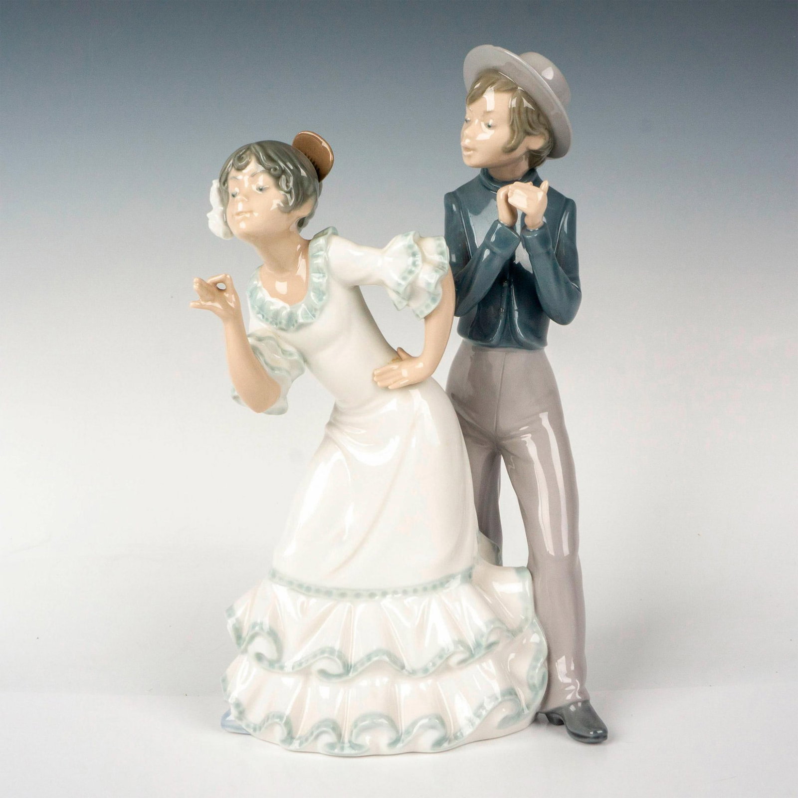 Nao by Lladro Figurine, Cantares Flamenco Dancers: A porcelain figurine depicting a couple side by side with the woman in a white dress and the man in a blue shirt, gray pants, and matching hat. Nao by Lladro backstamp. Dimensions: 6"L x 4.5"W x 11"H 