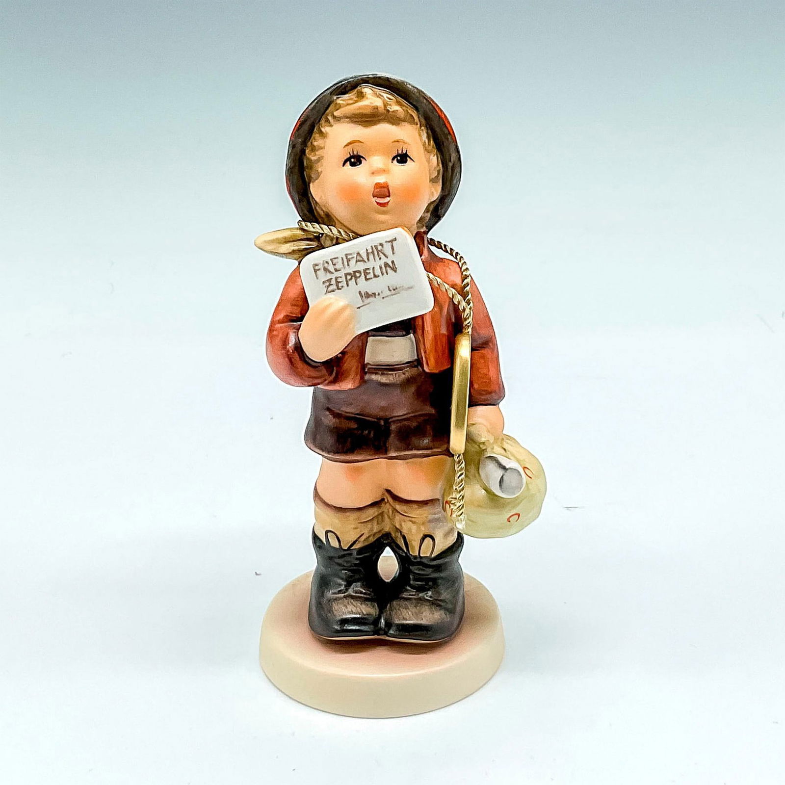 Goebel Hummel Porcelain Figurine, A Free Flight: Hand painted newspaper boy with headlines of the Zeppelin. Goebel backstamp. M.I. Hummel impressed mark. 569. This item has its original box: 5.5"L x 4"W x 4"H. Certificate of Authenticity included. I