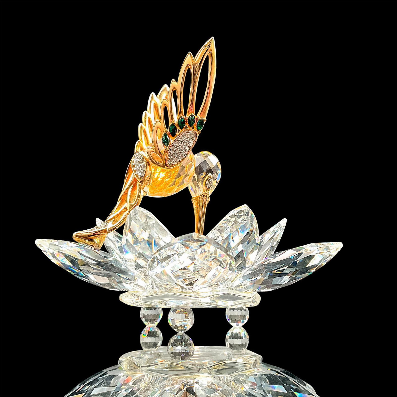 Swarovski Crystal Figurine, In Flight Hummingbird: Stunning faceted crystal figurine modeled as a hummingbird with gold-tone wings and beak studded with colorless and green Swarovski crystal gems. The hummingbird is featured pollinating a faceted crys