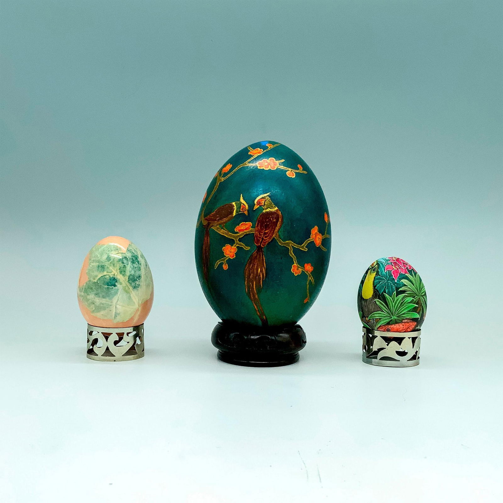 3pc Decorative Hand Painted Eggs with Bases: A set of three decorative eggs with fauna and foliage designs painted beautifully onto each item. One is a large green egg with birds atop a blossoming tree, one is painted in pastel colors with a lea