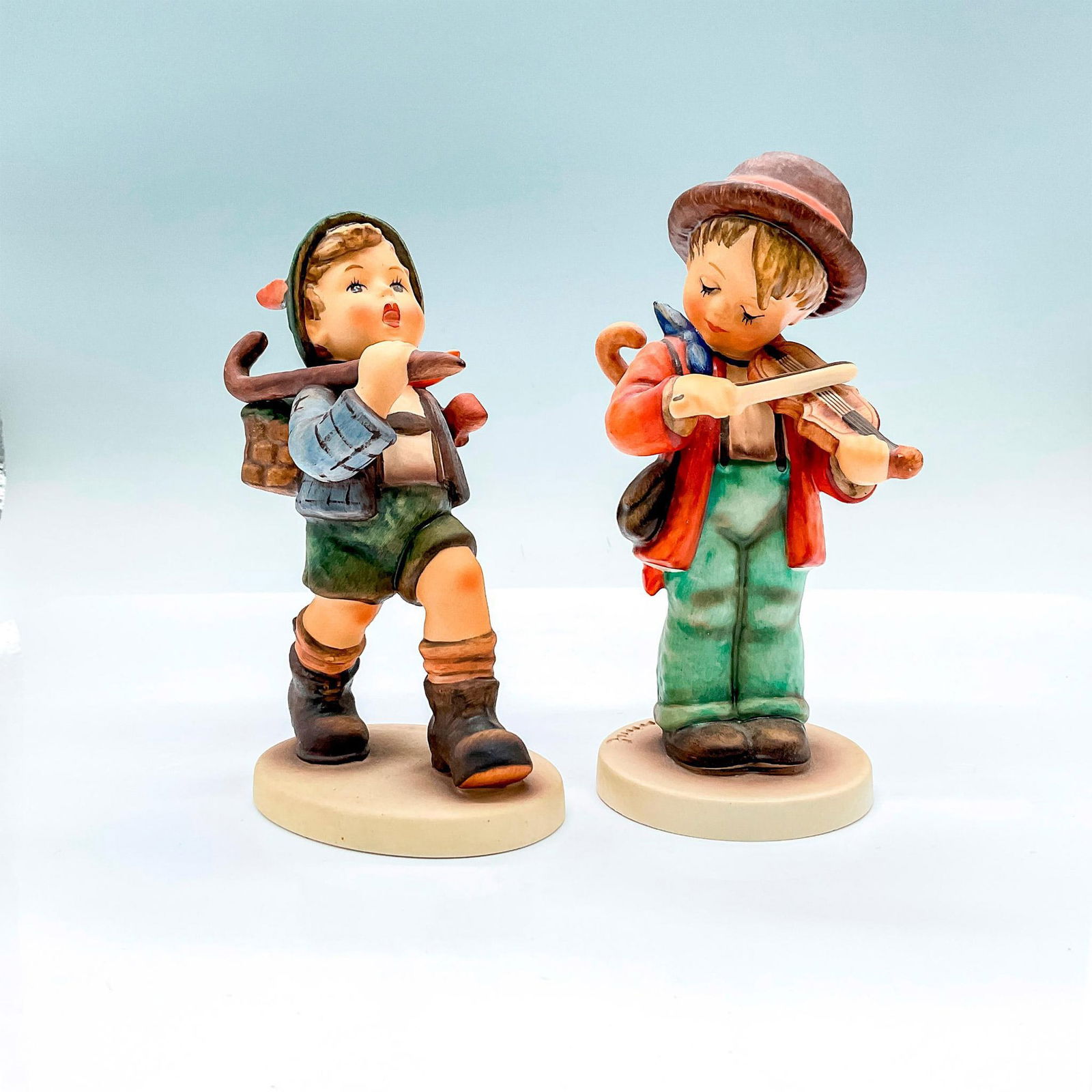 2pc Goebel Hummel Figurines, The Run A-Way + Little Fiddler: Hand painted porcelain figures. The Run A-Way (HUM 327) a young boy with a cane and basket of flowers. Little Fiddler (HUM 2/0) a young boy serenely playing a string instrument. Goebel backstamp. The