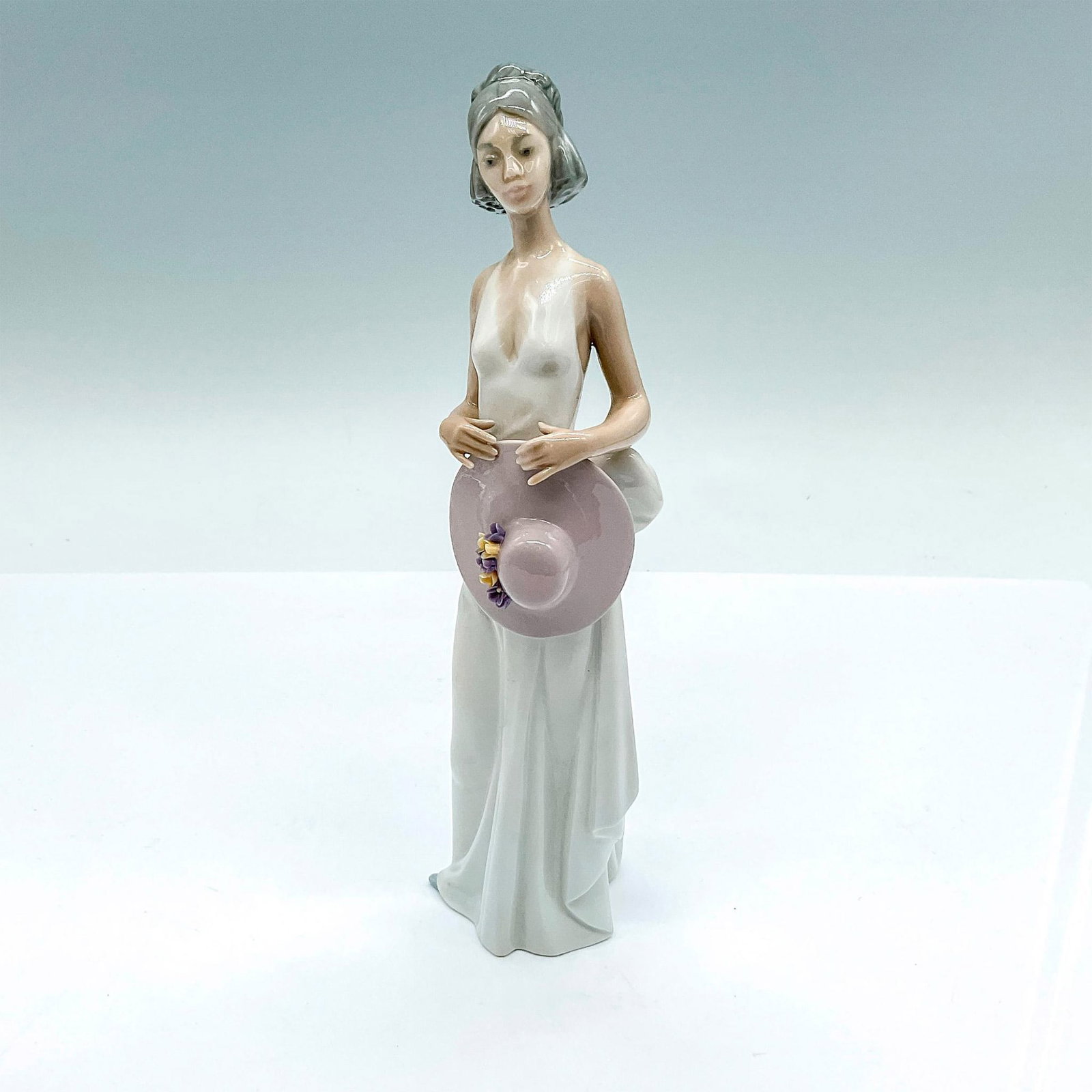 Summer Soiree 1005597 - Lladro Porcelain Figurine: A glossy figure of a young woman in long summer dress, clutching a lilac white brim hat with flowers. Lladro backstamp. Artist: Jose Puche Issued: 1989-1998 Dimensions: 3.25"L x 2.5"W x 8.25"H Manufac
