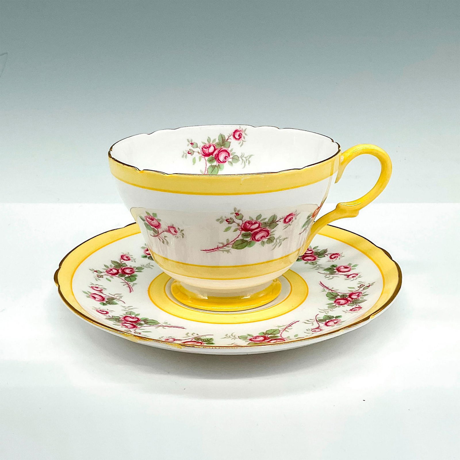 2pc Shelley Bone China Tea Cup and Saucer, Pink Roses: White bone china tea cup and saucer set with yellow bands, gilded gold trim, and revolving pink rose registers. Shelley backstamps. Tea cup: 4.5"L x 3.75"W x 2.5"H; saucer: 5.75" dia. Manufacturer: Sh