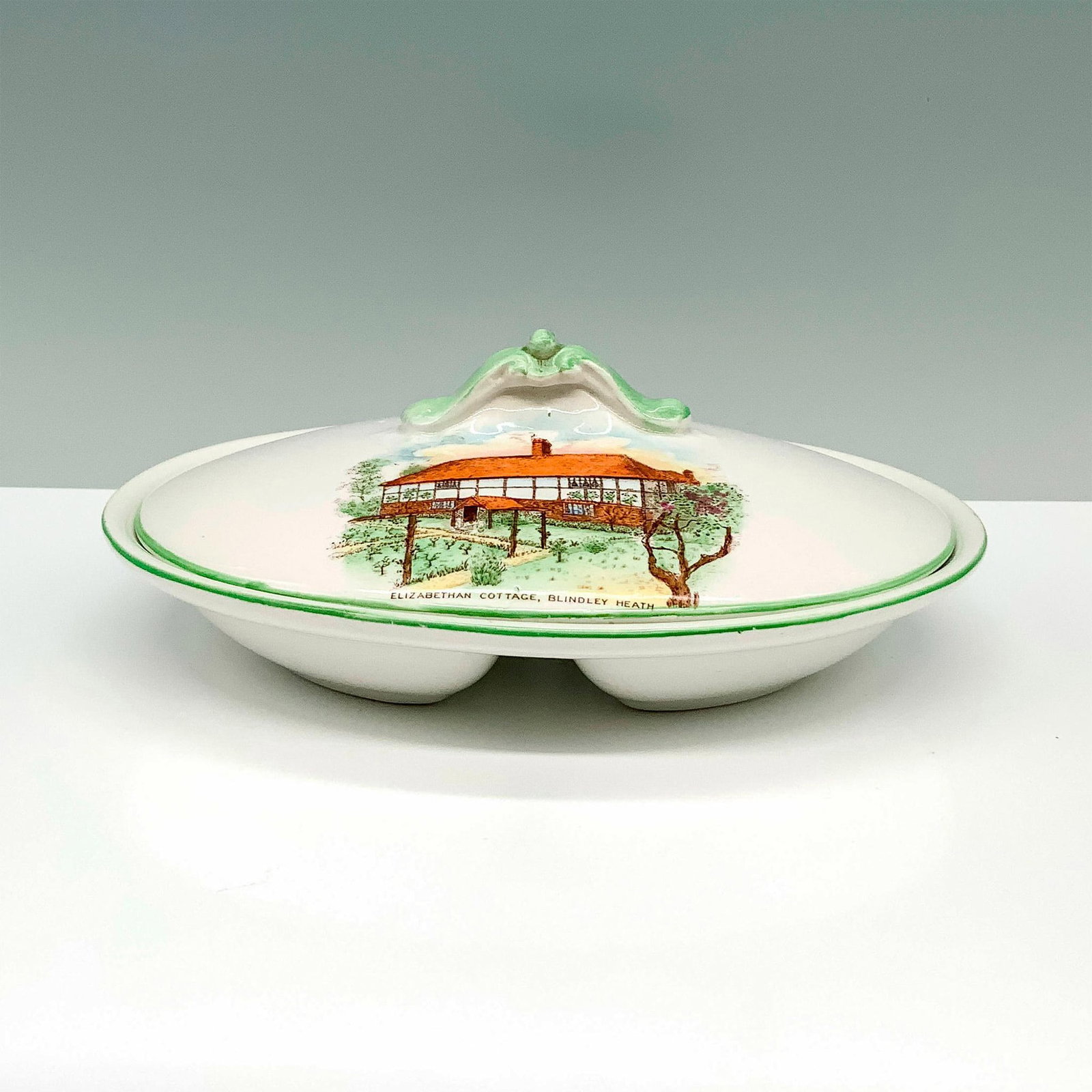 Shelley Bone China Divided Bowl and Lid, Elizabethan Cottage: A white bowl that has a raised center to divide the bowl in two along with a lid that has painting of a countryside cottage and its yard. Shelley backstamp. Dimensions: 8"L x 6"W x 3.25"H Manufacturer