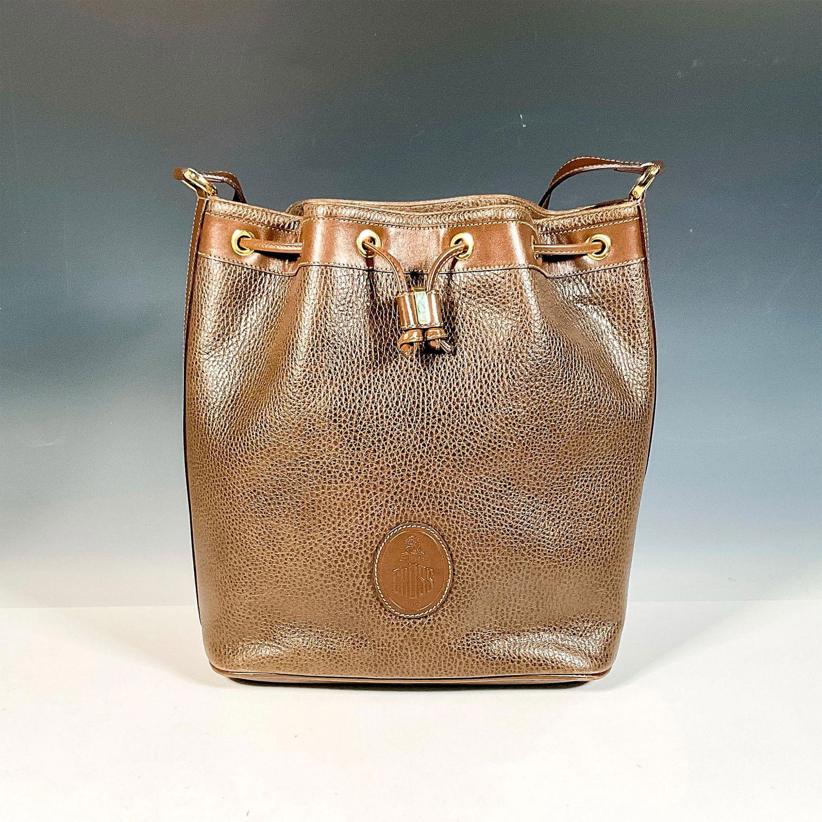 Mark Cross Brown Leather Handbag: Brown bucket style handbag with drawstring closure and adjustable shoulder strap. Mark Cross embossed logo on the front. Includes original box, 12"L x 10.75"W x 4.75"H. Dimensions: 11.5"L x 4.75"W x 1