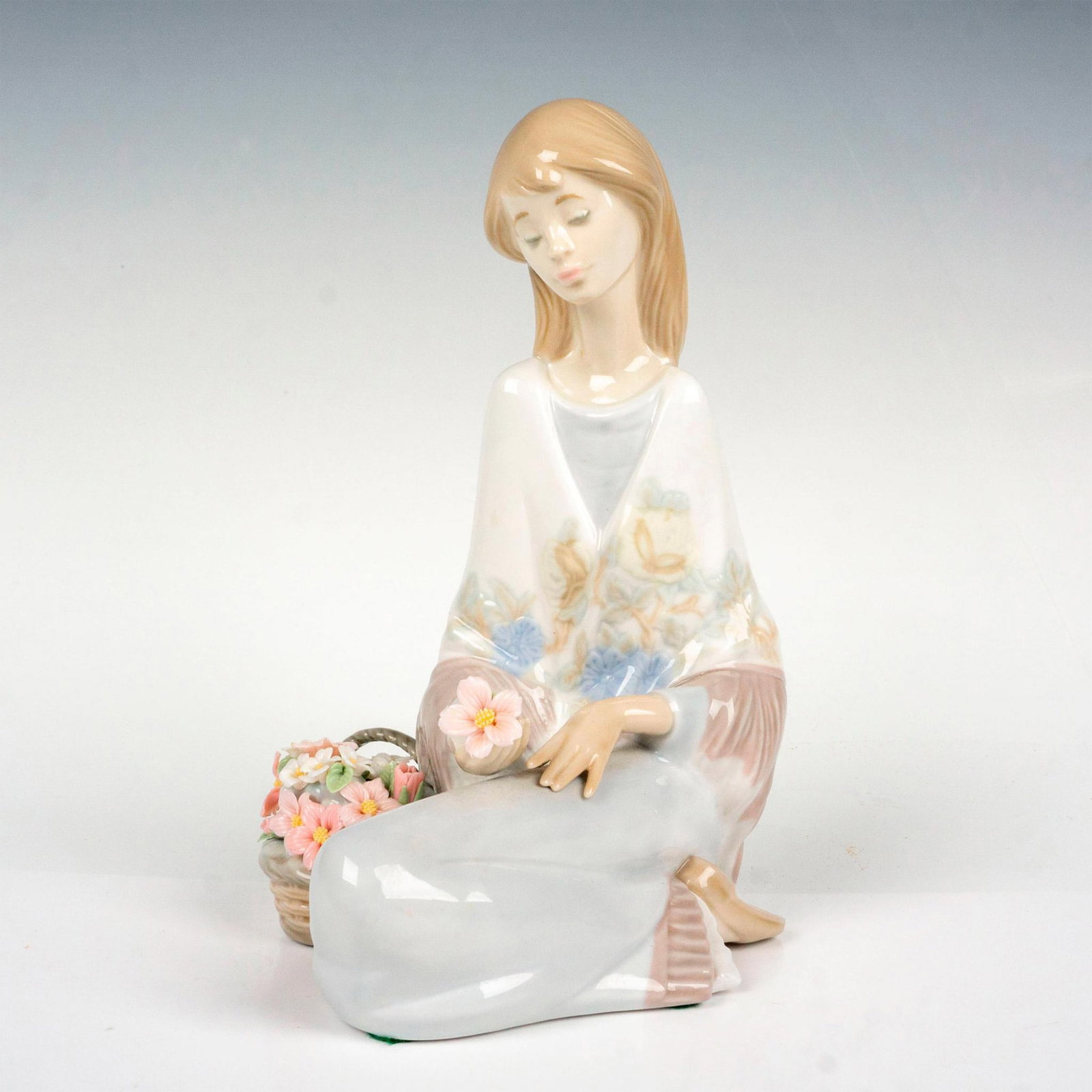 Flower Song 1007607 - Lladro Figurine: Young woman seated next to a basket of flowers. Lladro backstamp. This item has its original box which measures: 10"L x 9"W x 6.5"H. Artist: Juan Huerta Issued: 1989 Dimensions: 5.5"L x 4.5"W x 7.25"H