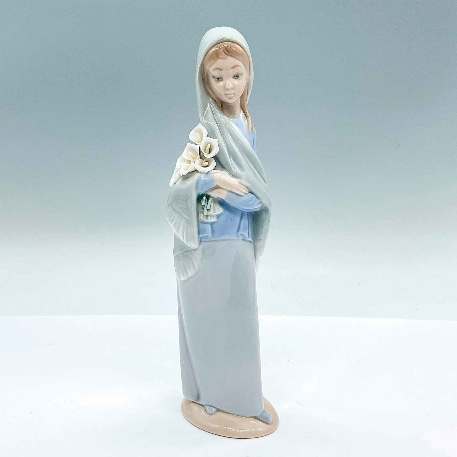Girl With Calla Lilies 1004650 - Lladro Porcelain Figurine: A glossy figure of a woman in a blue shawl embracing a bouquet of lilies. Lladro backstamp. Artist: Vincente Martinez Issued: c. 1969-1998 Dimensions: 2.5"L x 2.25"W x 9"H Manufacturer: Lladro Countr