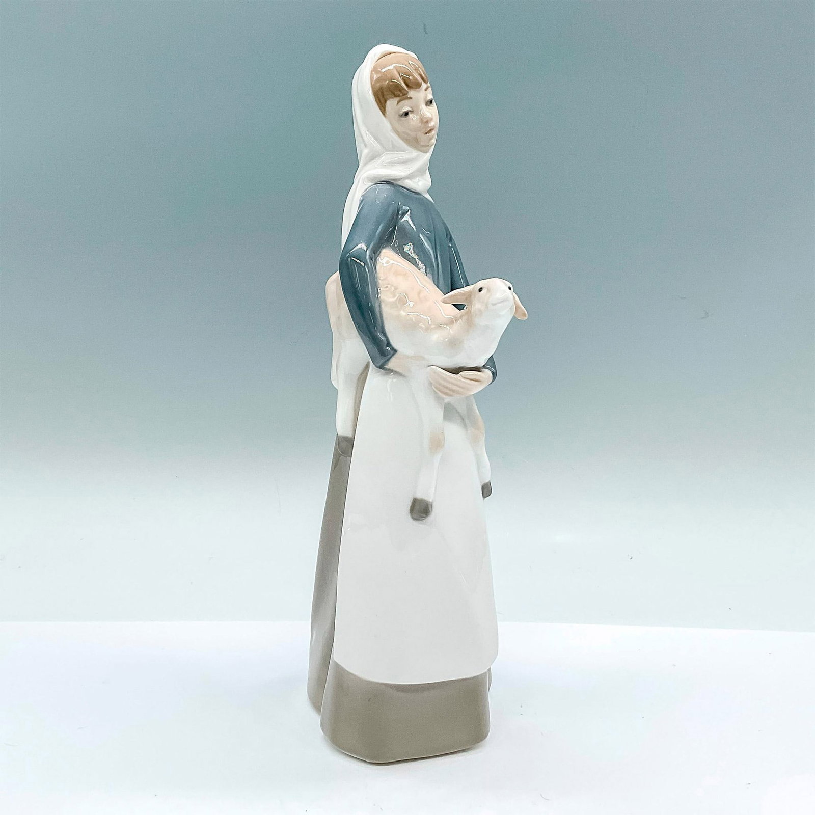 Girl With Lamb 1004584 - Lladro Porcelain Figurine: A glossy figure of a young girl holding a lamb in her arms. Lladro backstamp. Artist: Alfredo Ruiz Issued: c. 1969-1993 Dimensions: 4.5"L x 3.5"W x 10.75"H Manufacturer: Lladro Country of Origin: