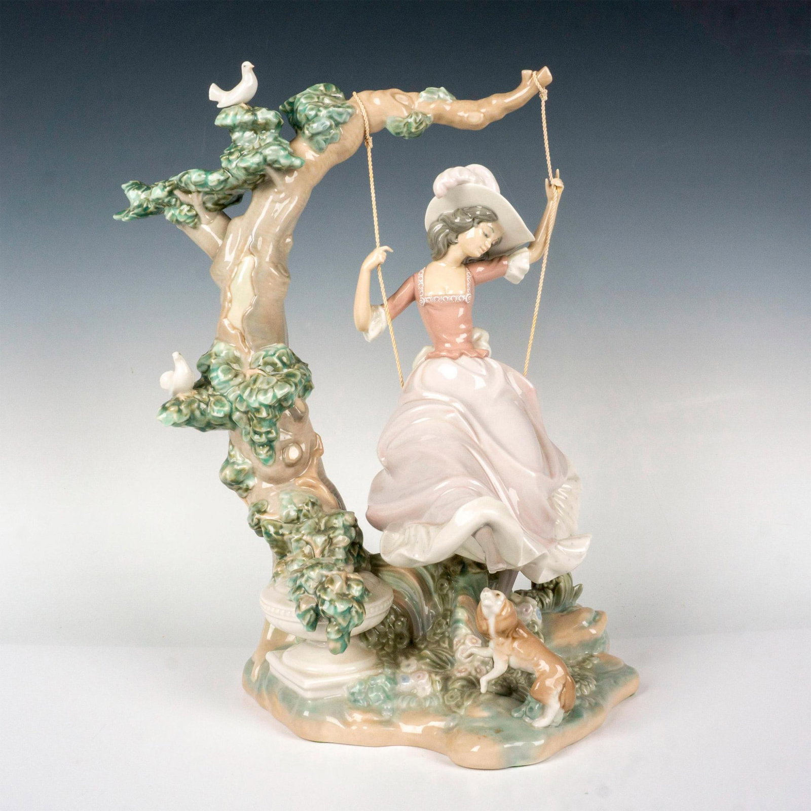 Victorian Girl on Swing 1001297 - Lladro Figurine: Composed of glossy glazed porcelain depicting a woman on a swing with a large tree, bird, and dog motifs surrounding her. Lladro backstamp. Artist: Salvador Debon Issued: 1974-1989 Dimensions: 13"L x 