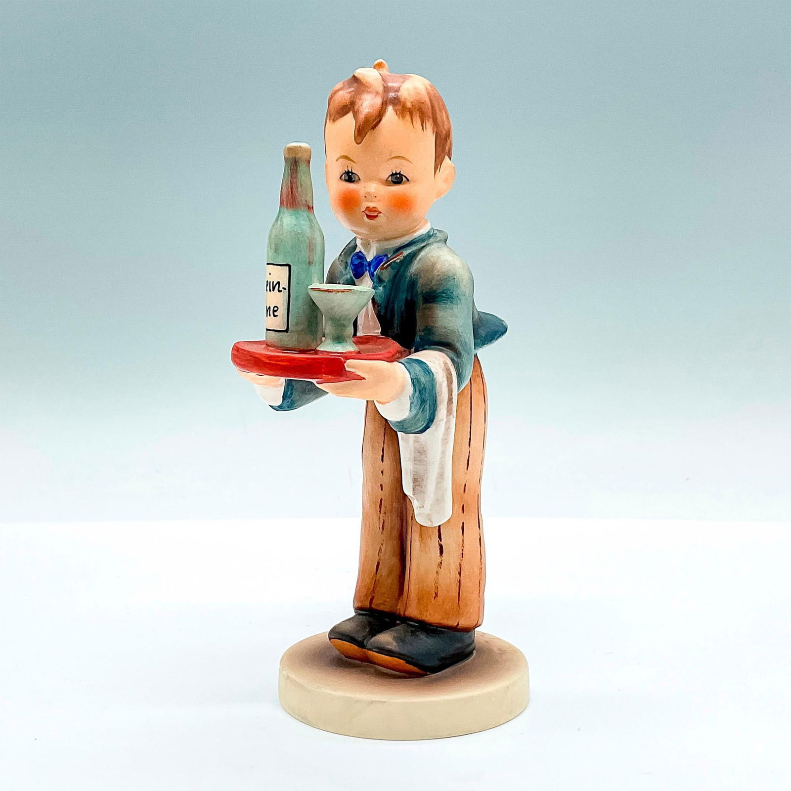 Goebel Hummel Porcelain Figurine, Waiter HUM 154: Hand painted figure of a young man serving a bottle wine. Goebel backstamp. Artist: M.I. Hummel Issued: c. 1972-1979 Dimensions: 3.25"L x 2.5"W x 6"H Manufacturer: Goebel Hummel