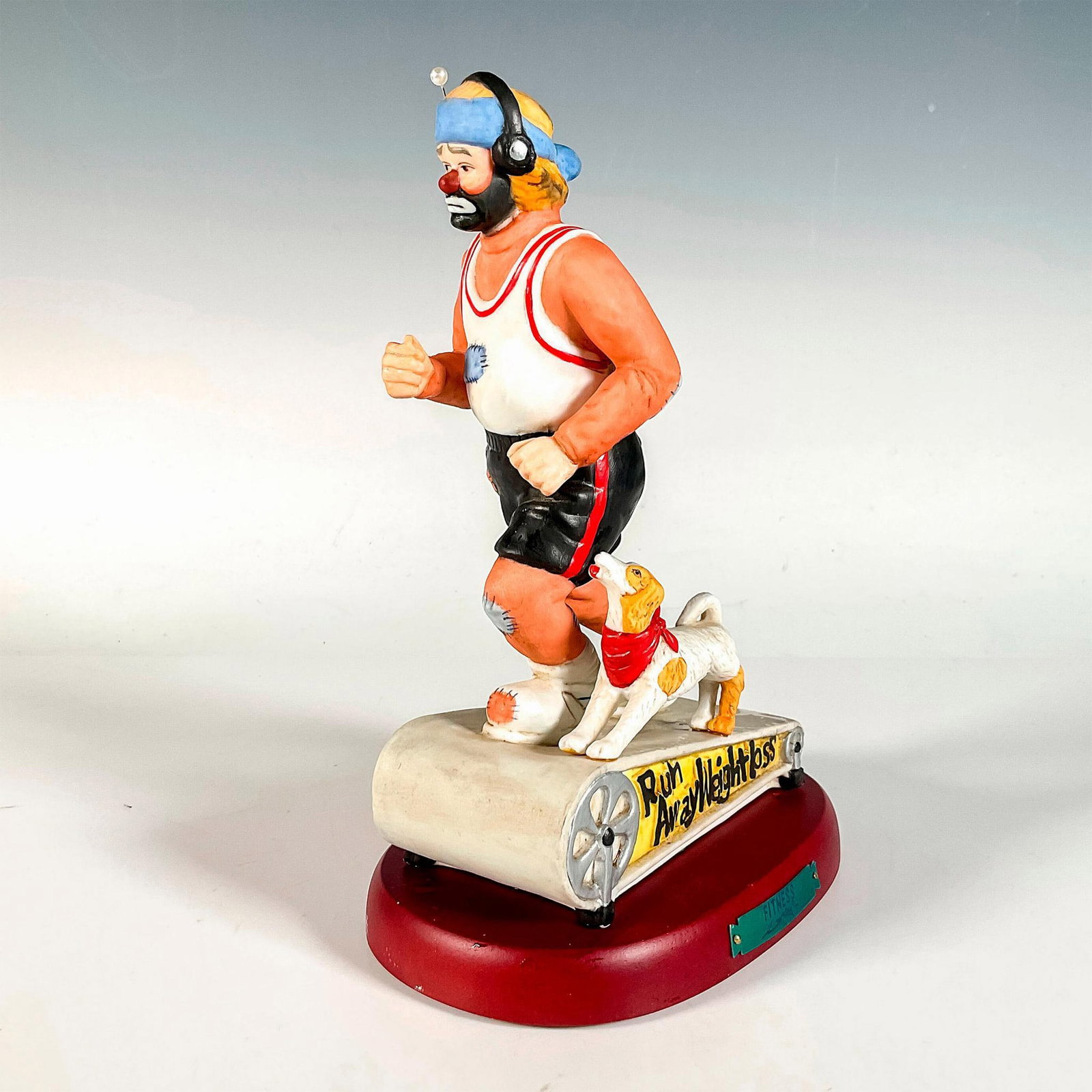 Flambro Emmett Kelly Jr. Figurine, Fitness: Hand painted bisque porcelain depicting figural clown Weary Willie running on the treadmill with his dog. Rests on a wooden base with gold-tone titled plaque. Emmett Kelly Jr. signature backstamp. Fla