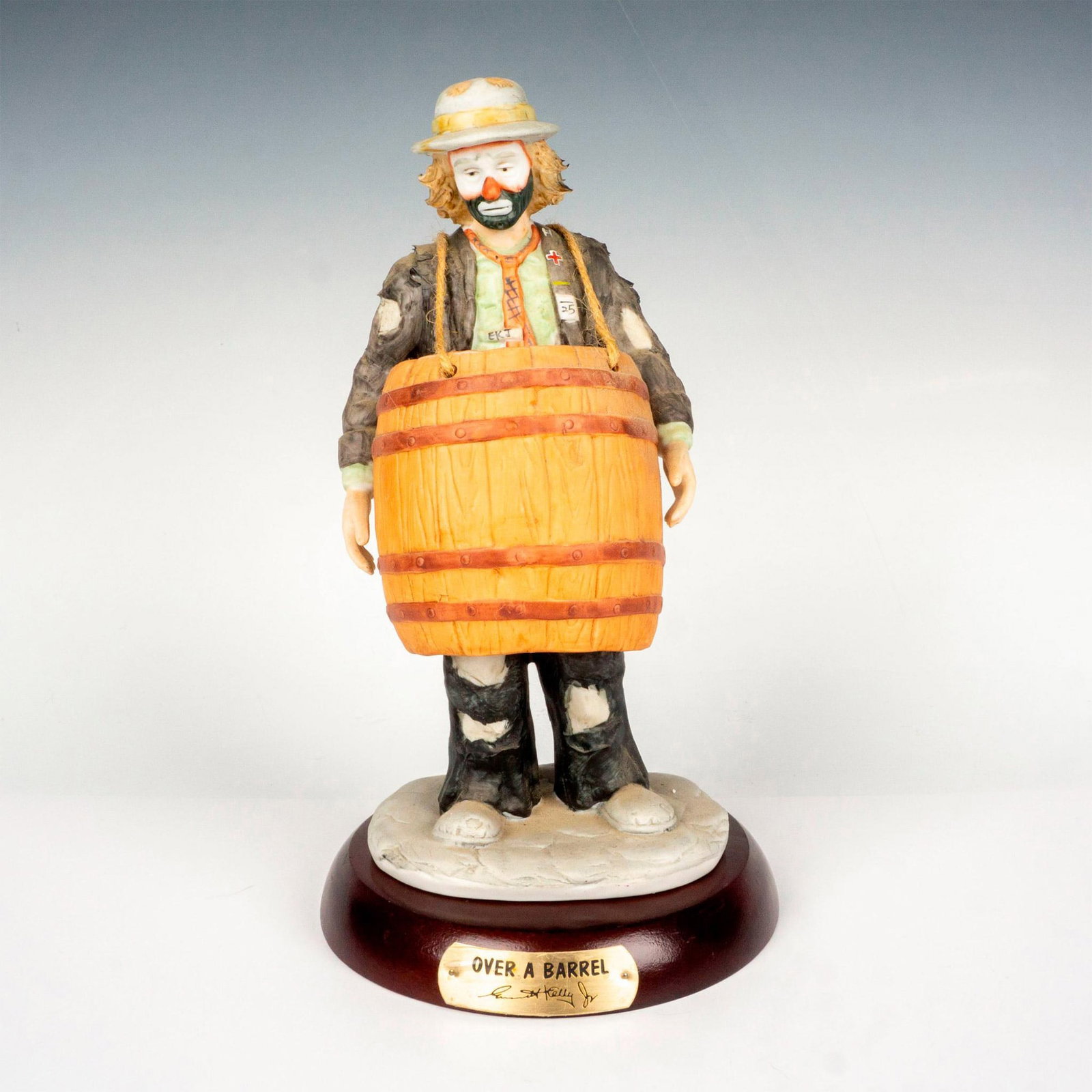 Flambro Emmett Kelly Jr. Figurine, Over A Barrel + Base: Bisque porcelain figural of the character Weary Willie standing with a barrel around him. Flambro Imports stamp. This item has a base included: 1"H x 6"dia. This item has its original box: 8.5"L x 8.2