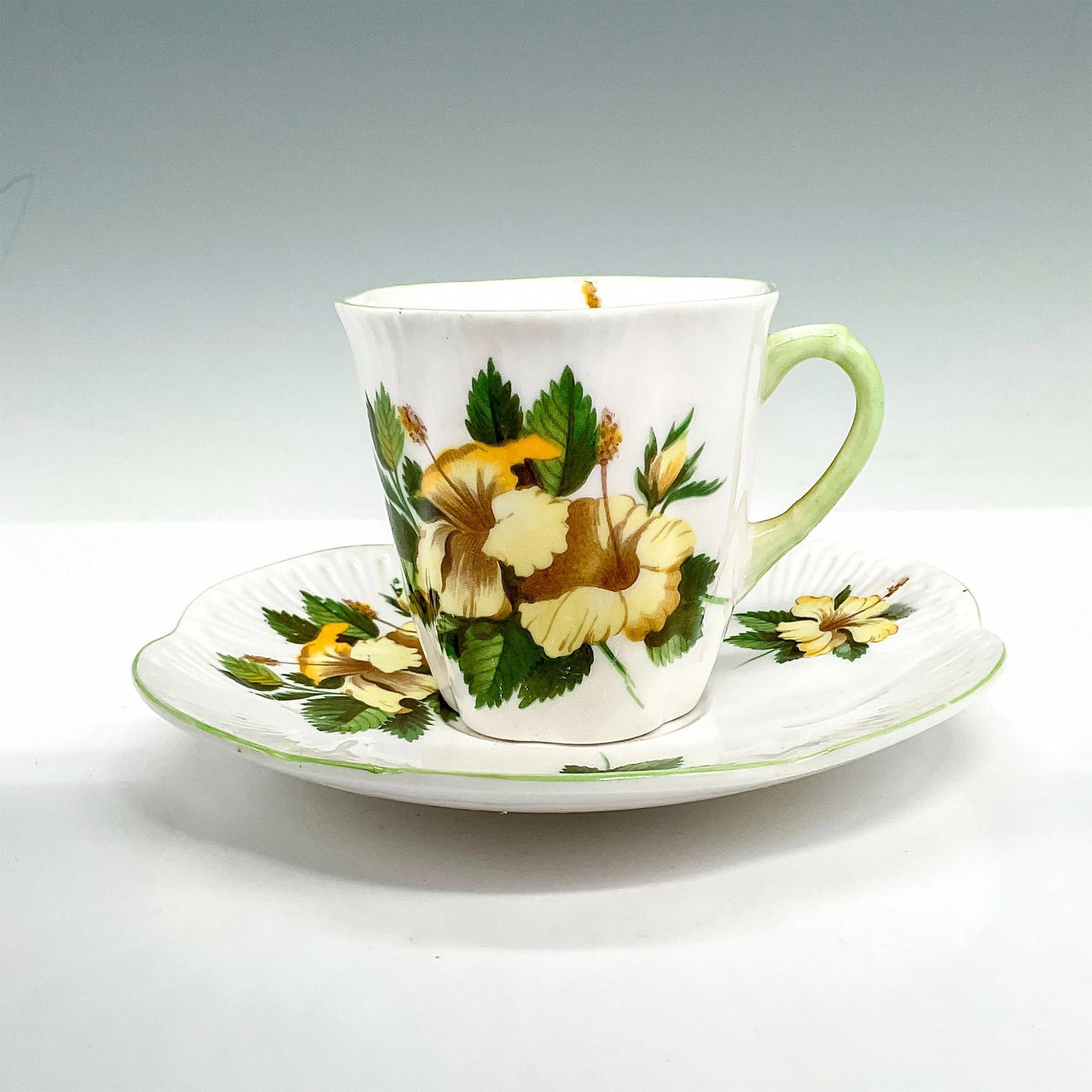 Shelley China Demitasse Cup and Saucer, Hibiscus: White bone china tea cup and saucer set with cream-yellow hibiscus designs, scalloped edges, and green trim. Shelley backstamps. Cup: 3"L x 2.5"W x 2.5"H; saucer: 4.75" dia. Manufacturer: Shelley Coun