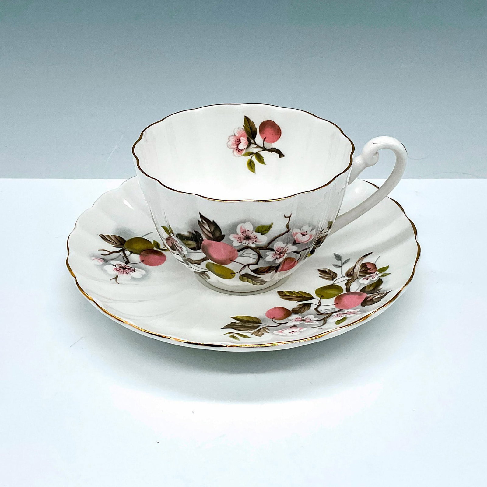 Shelley China Teacup and Saucer Set, Pink & Green Cherry: Scalloped teacup and matching saucer set of fine white china decorated with pink and green cherry blossoms finished with gold trim. Shelley backstamps. Cup: 4.5"L x 3.5"W x 2.5"H. Saucer: 1"H x 5.75"d