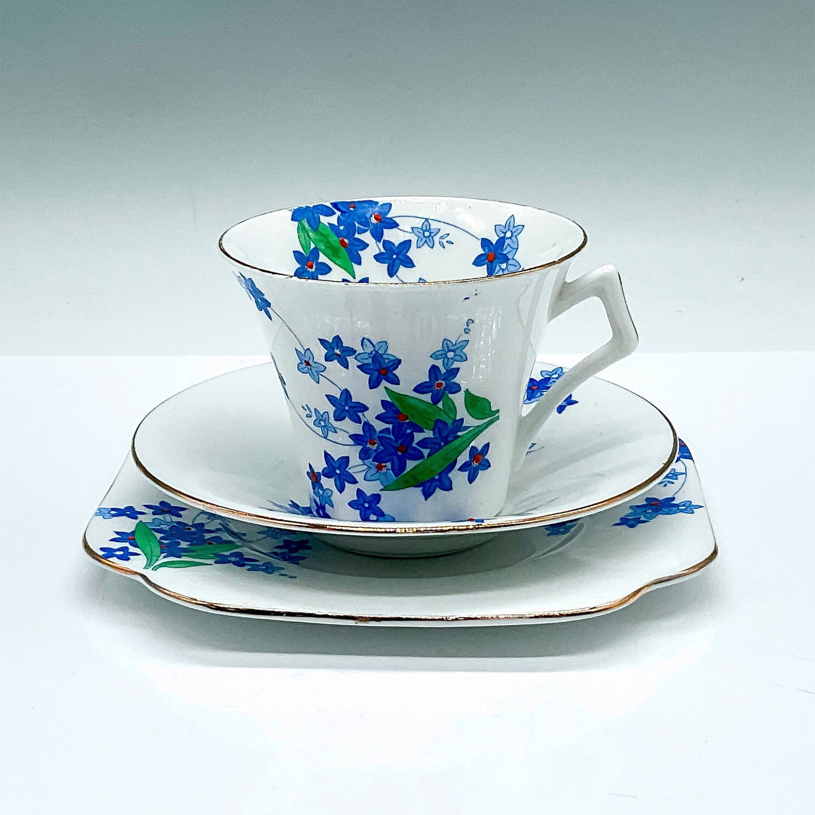 Colclough Bone China Blue Floral Tea Trio No. 4117: White cup, saucer, and dessert plate. Decorated with blue star-shaped flowers and gilded trims. Colclough backstamps. Cup: 4"L x 3.25"W x 2.75"H. Saucer: 5.25"W x 1"H. Dessert plate: 6"L x 6"W x 0.5"H