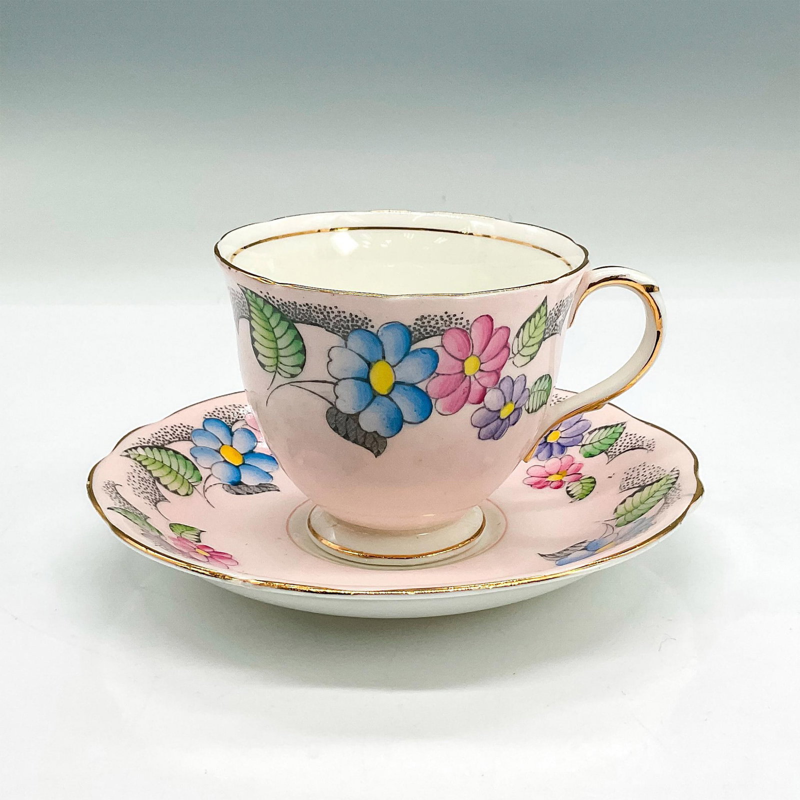 Foley China Cup and Saucer, Pink with Colorful Flowers: Bone china tea cup and saucer in pale pink with blue, purple and dark pink floral design; gilt rims. Foley China backstamp. Cup 4"L x 3.25"W x 2.75"H. Saucer 0.75"H x 5.5" dia. Manufacturer: Foley Chi