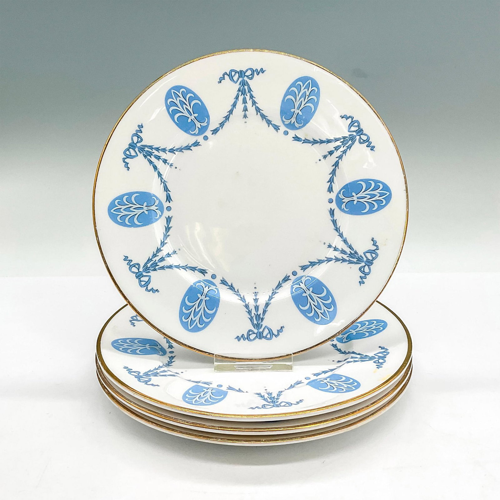 4pc Shelley England Plates, Osterley: Four bone china plates with a blue bow, floral chain and oval insignia style design, gilt rims. Shelley England backstamp. Dimensions: 0.5"H x 6.5"dia Manufacturer: Shelley Country of Origin: England