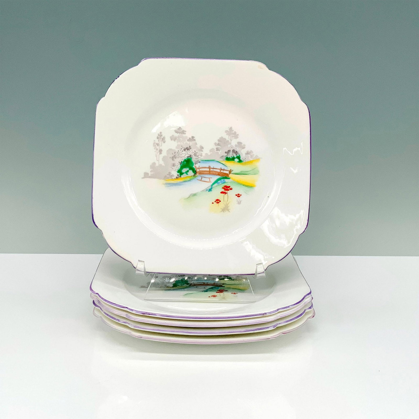 5pc Shelley Bone China Quaint Trail Plates: A set of 5 white plates with lavender-colored borders that depict a woodland scene with a river and wooden bridge to cross. Shelley backstamp. Dimensions: 6.25"L x 6.25"W x 0.25"H Manufacturer: Shelle
