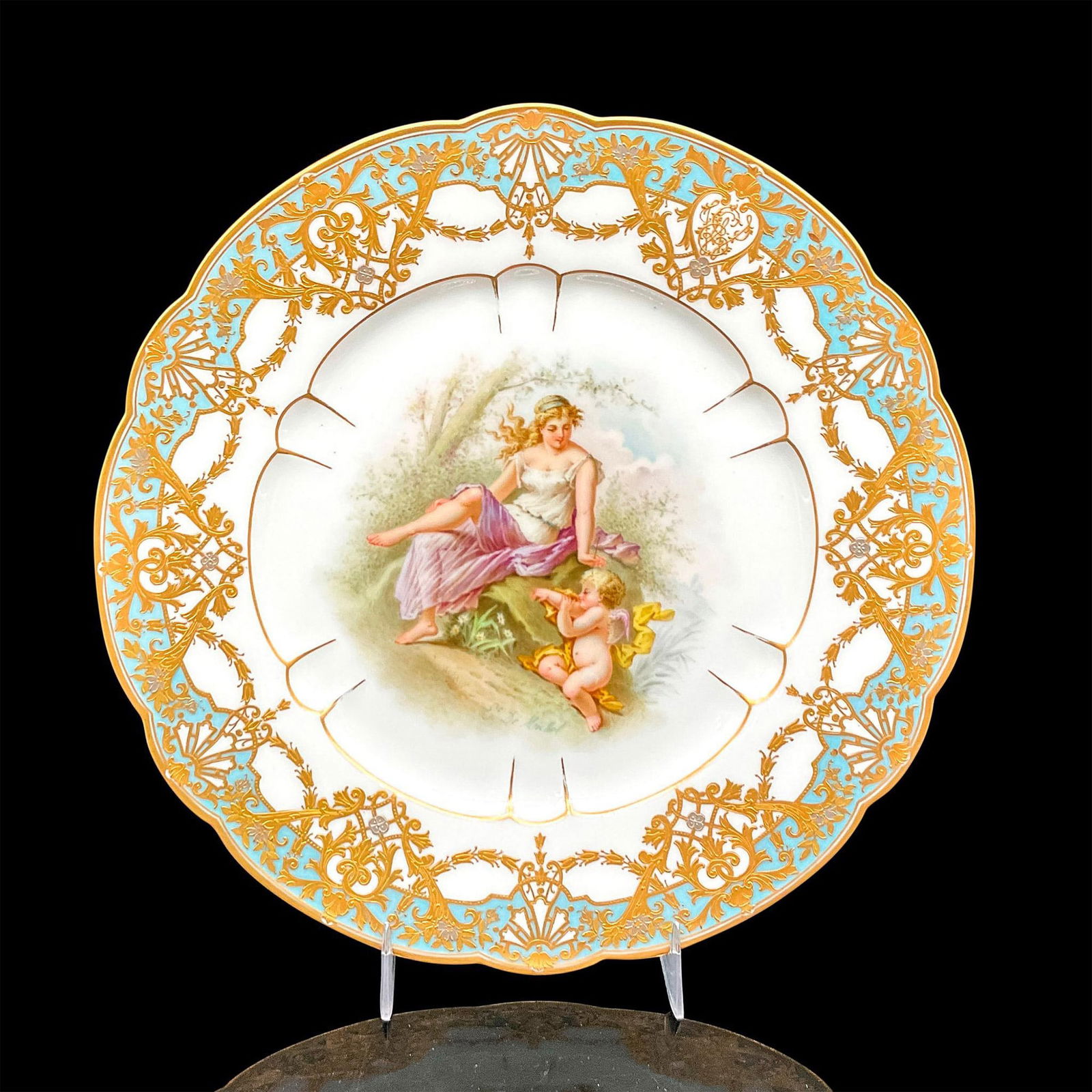 Sevres Porcelain Plate, Nude Lady in The Garden: Hand painted porcelain decorative plate with an image of a lady in a white and purple dress sitting on a rock next to a small cherub. Ornate gold design on lip. Sevres mark backstamp. Artist signed. D