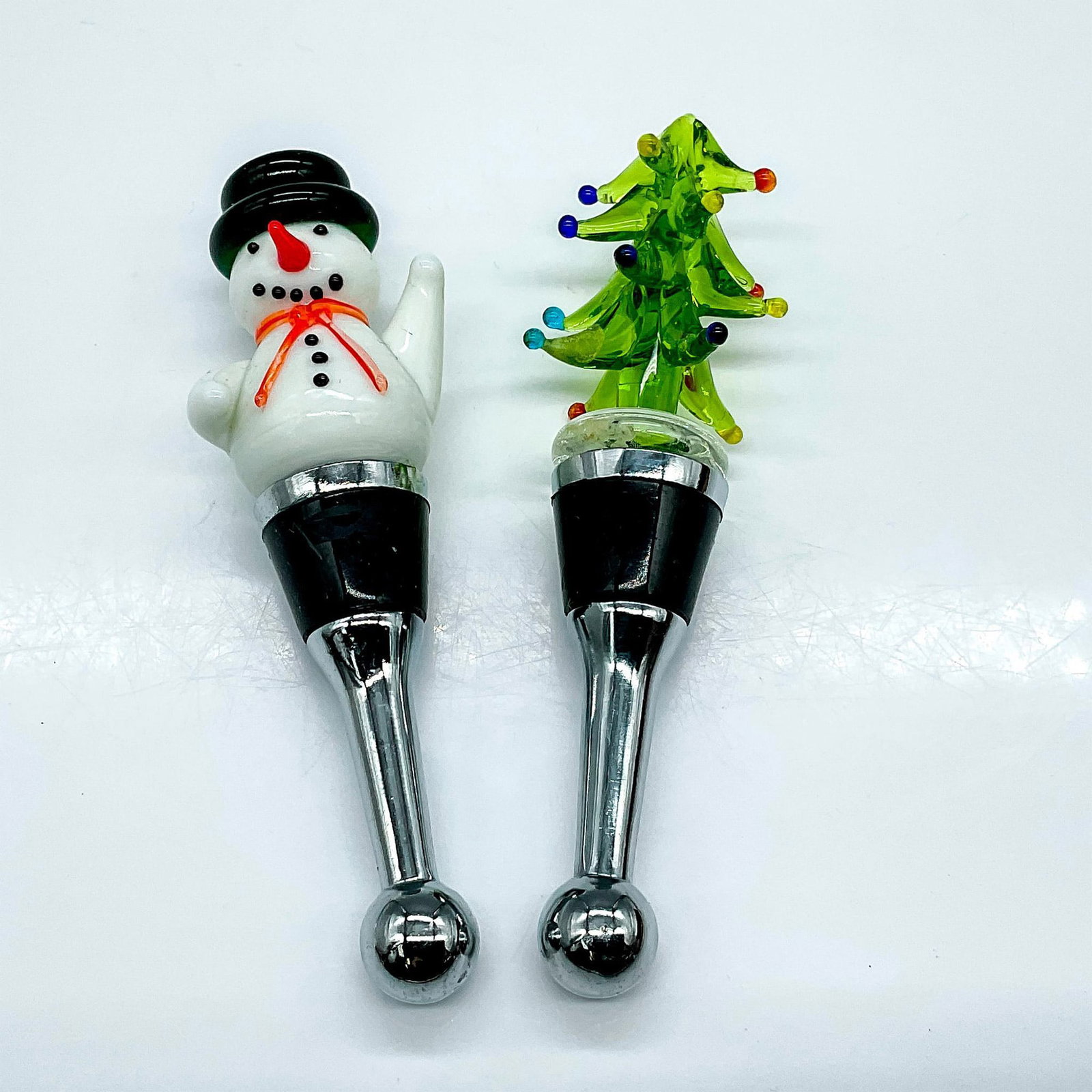 2pc Christmas Art Glass Wine Stoppers: This festive pair of holiday stoppers, constructed with metal and rubber for secure sealing of wine or spirits bottles, showcases handcrafted art glass tops portraying an endearing snowman and a cheer