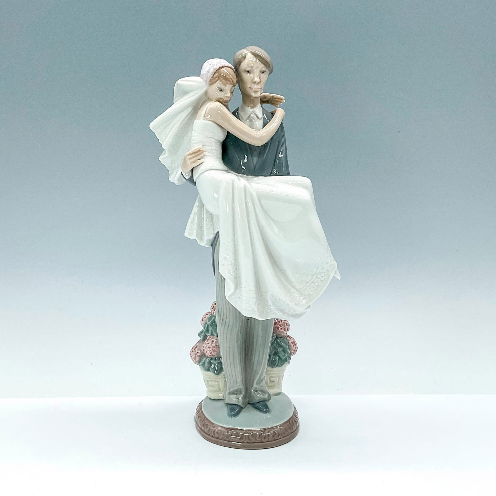 Over the Threshold 1005282 - Lladro Porcelain Figurine: This glazed figurine beautifully captures the moment of a bride and groom walking over the threshold. Lladro backstamp. Artist: Juan Huerta Issued: 1985-2007 Dimensions: 5.5"L x 3.75"W x 10.25"H Manuf