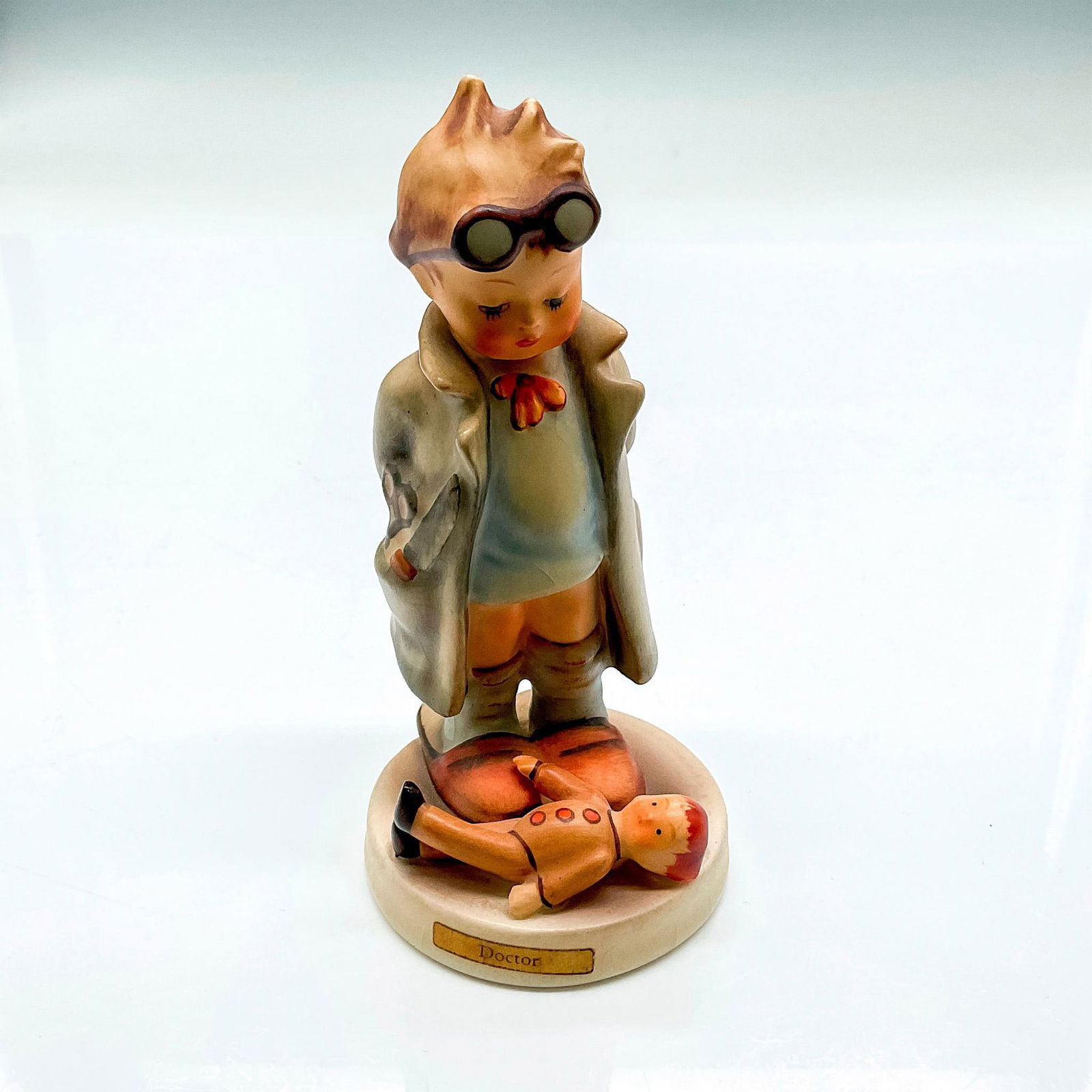 Goebel Hummel Porcelain Figurine, Doctor: Hand painted boy with goggles and lab coat overlooking a doll patient. Hummel backstamp W Germany. Impressed mark 127. Issued: 20th century Dimensions: 4.75"H x 2.25" dia. Manufacturer: Goebel Hummel