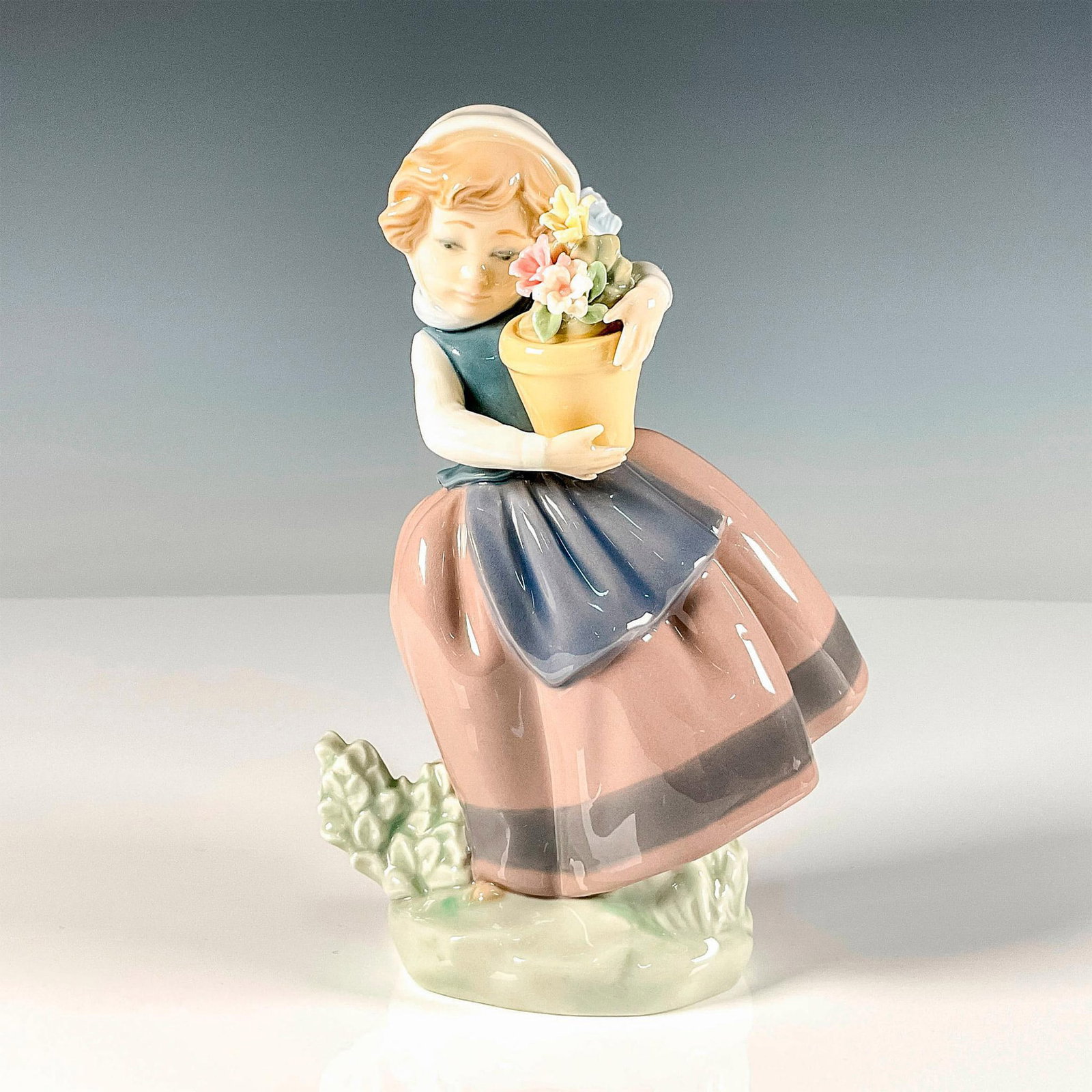 Spring Is Here 1005223 - Lladro Porcelain Figurine: Glossy finish figure modeled as a young village girl holding a pot of fresh flowers as the wind blows. Lladro backstamp. Artist: Jose Puche Issued: 1984-2021 Dimensions: 4"L x 3.25"W x 6.75"H Manufact