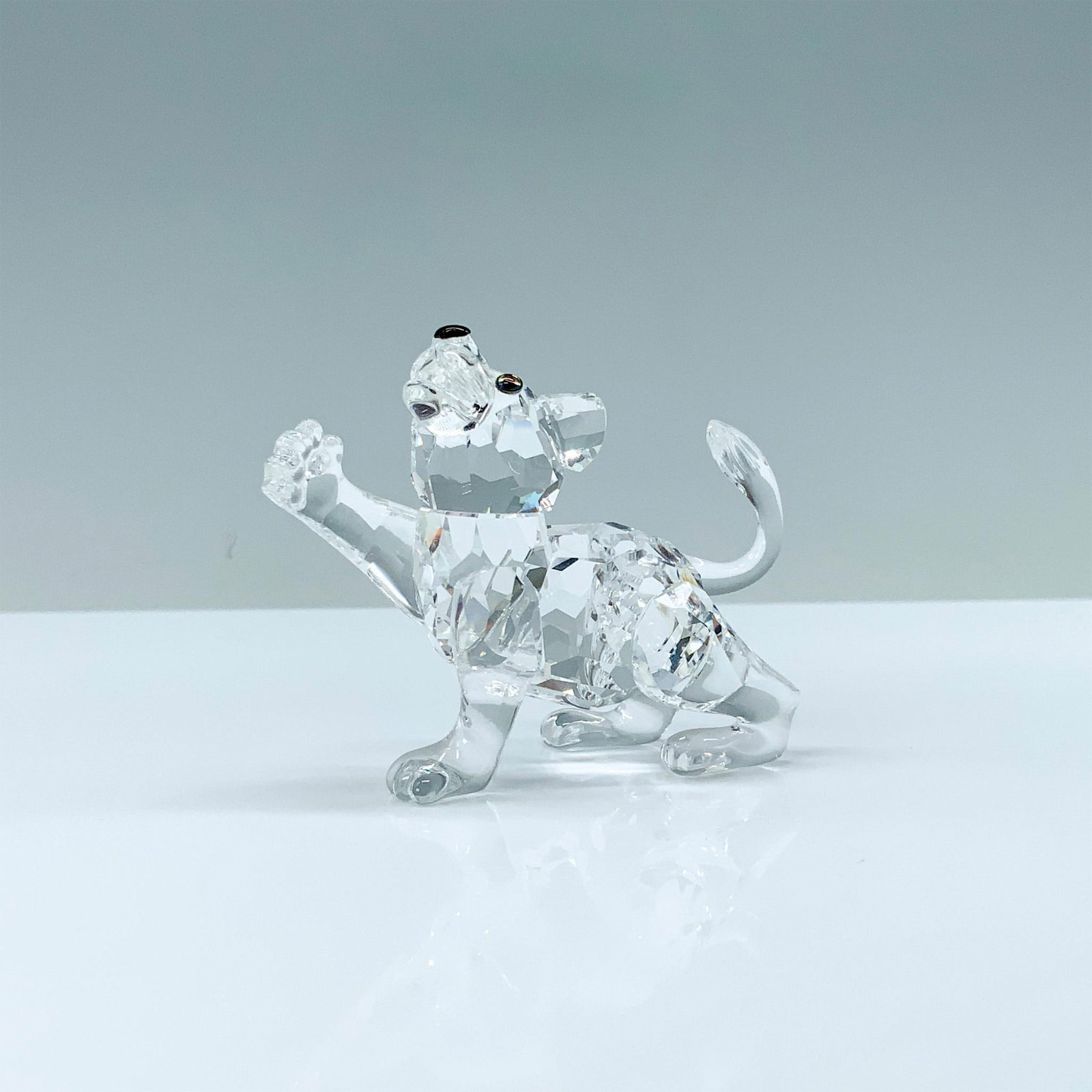 Swarovski Crystal Figurine, Lion Cub: A clear crystal figure of a young lion stretching his paw outward with a raised tail and melon-colored crystals for eyes. Swarovski backstamp. This item has its original box. Dimensions of the origina